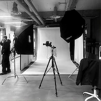 A black and white photo of an empty e-commerce photography set at LRP studios, featuring a seamless white backdrop, a camera on a tripod, and large studio lights.