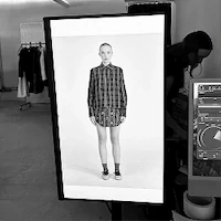 A black and white behind-the-scenes photo of e-commerce photography at LRP studios, focusing on a large vertical monitor displaying an image of a model, with a crew member working at a digital station in the background.