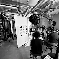 A black and white behind-the-scenes photo of e-commerce photography at LRP studios, showing crew members seated on set facing a model posing on a white backdrop, with a mood board and lighting equipment visible.