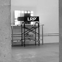 A black and white photo of a hair and makeup station prepared for e-commerce photography at LRP studios, featuring a tall director's chair with the LRP logo positioned in front of a lighted vanity mirror.