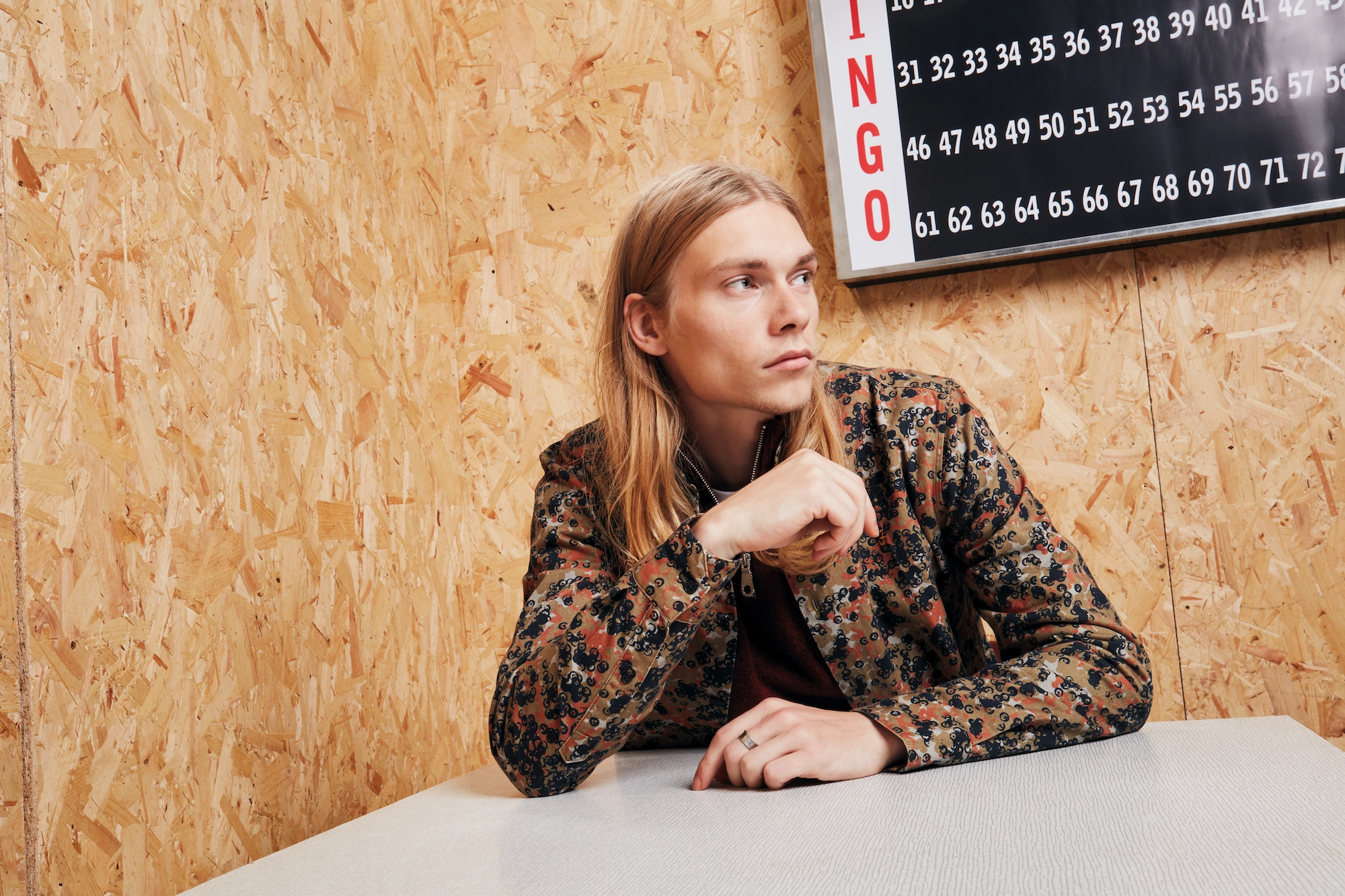 Male model sitting by the table with a intricate jacket and Bingo board behind him, posing for Farah Menswear creative shoot