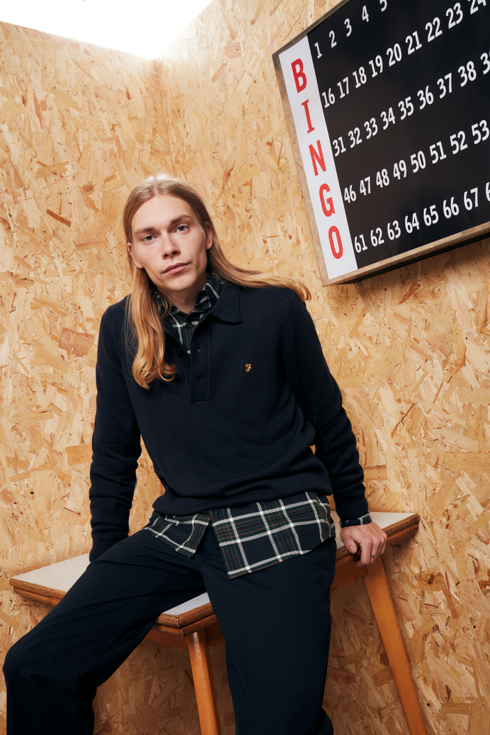 Male model posing whilst leaning on the table, wearing dark jumper with checkered short underneath and dark trousers. Shot for Farah by LRP Studios in London