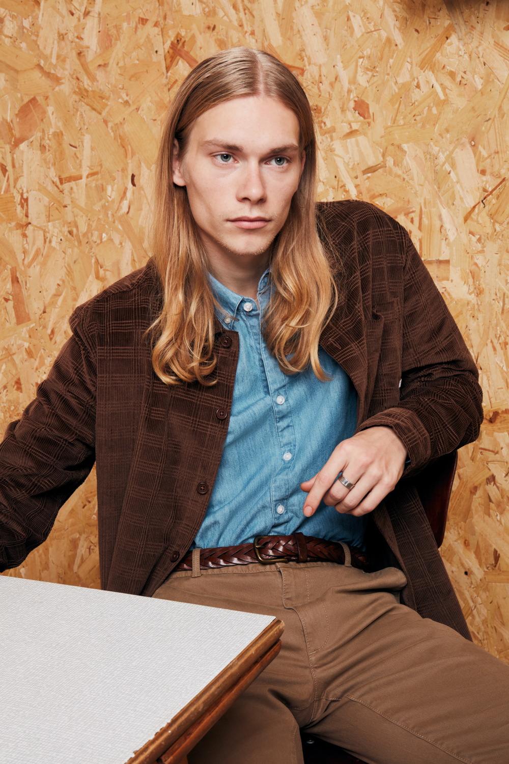 Male model with brown sweater and light blue shirt and beige trousers sitting by the table