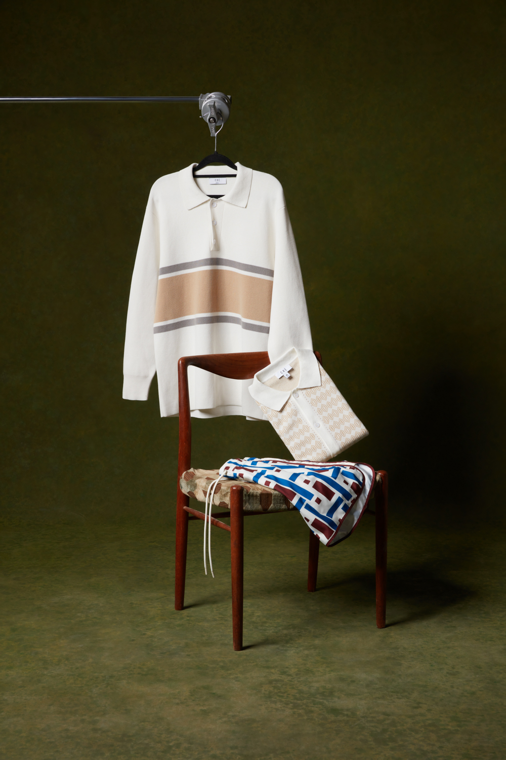 Che Studios men's fashion styling featuring a long-sleeve striped knit polo, a short-sleeve geometric patterned polo shirt, and retro geometric print shorts arranged on a vintage wooden chair