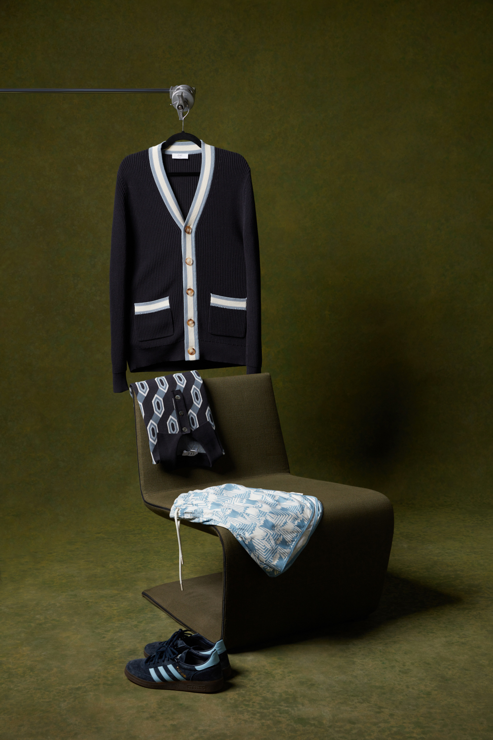 Che Studios men's fashion styling featuring a brown utility jacket layered over a patterned polo shirt, alongside folded cream waffle-knit and brown knit shirts arranged on a vintage wooden chair
