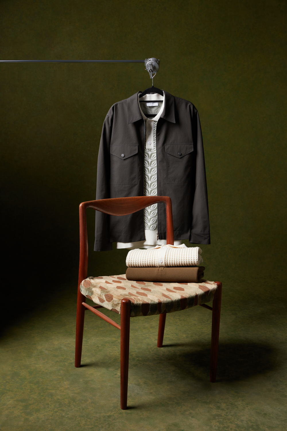 Che Studios men's fashion styling featuring a navy ribbed cardigan, geometric patterned knit shirt, and blue printed shorts, arranged around a green chair with navy suede Adidas sneakers