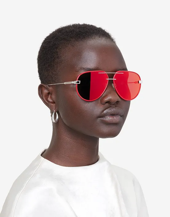 Woman with short hair wearing red Rimowa aviator sunglasses and silver hoop earrings against white background.