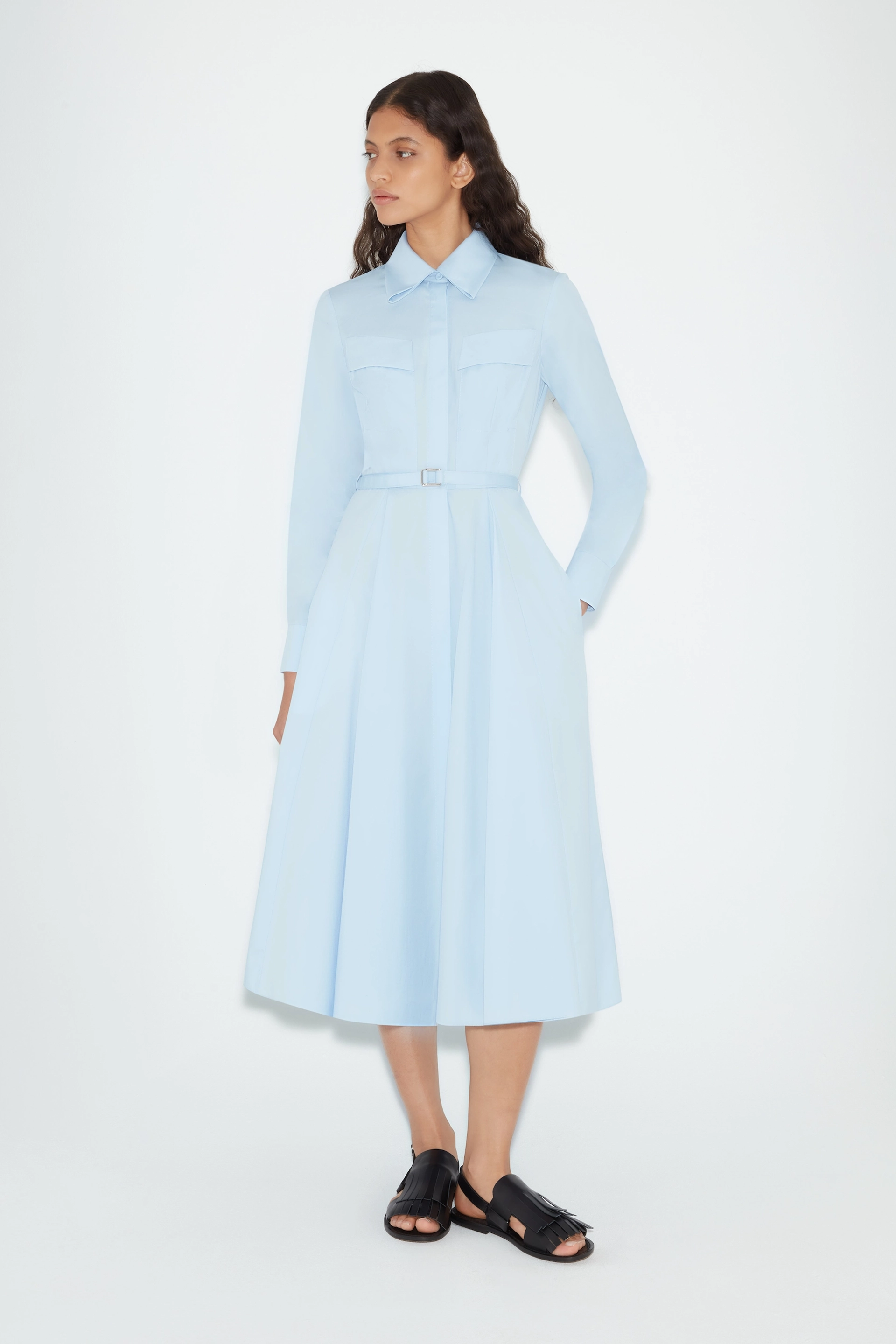 Woman wearing a light blue belted midi Emilia Wickstead dress with long sleeves and black open-toe sandals.