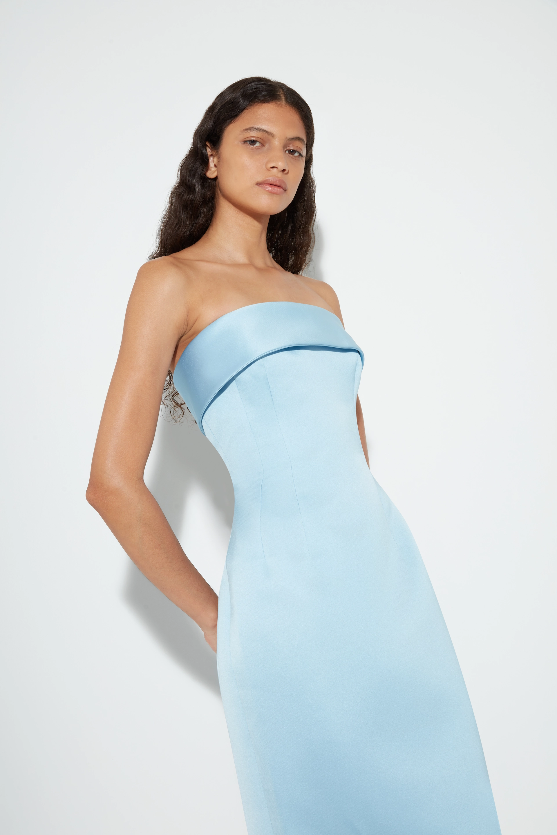 Woman with long wavy hair wearing a strapless light blue Emilia Wickstead fitted dress against a plain white background.