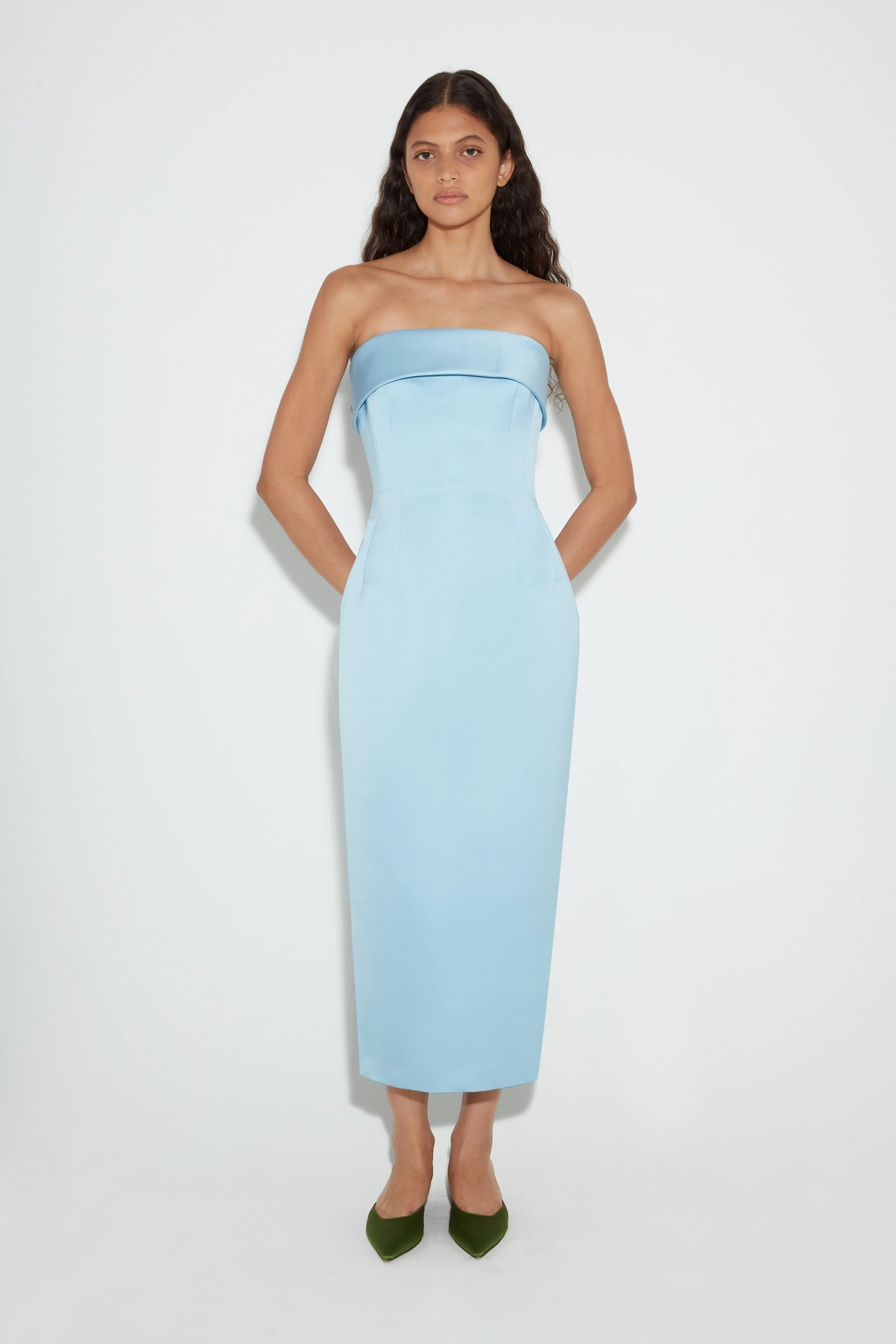 Woman with long dark hair wearing strapless light blue midi Emilia Wickstead dress and green pointed shoes.