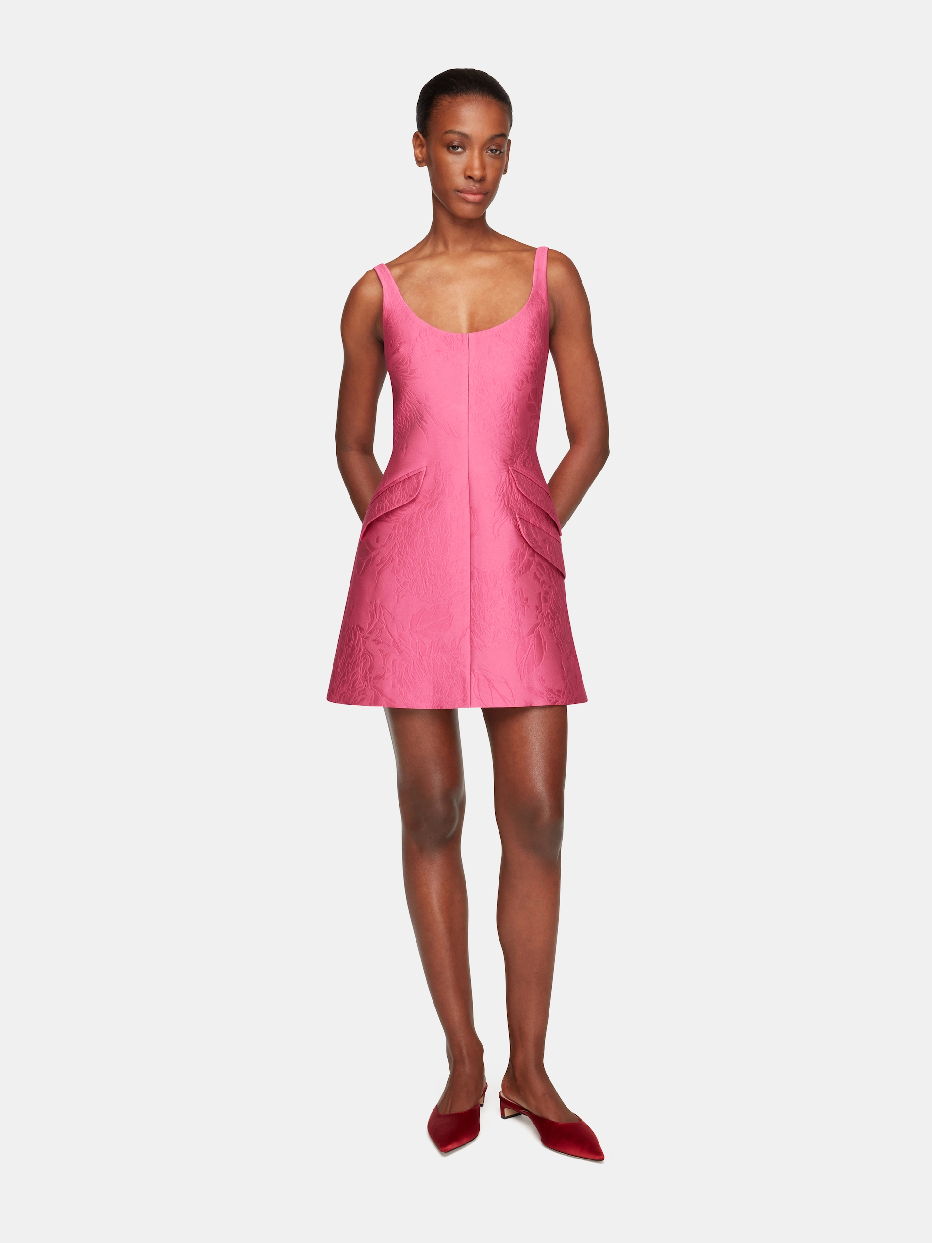 Woman in a sleeveless pink textured Emilia Wickstead dress with flap pockets wearing red pointed shoes.