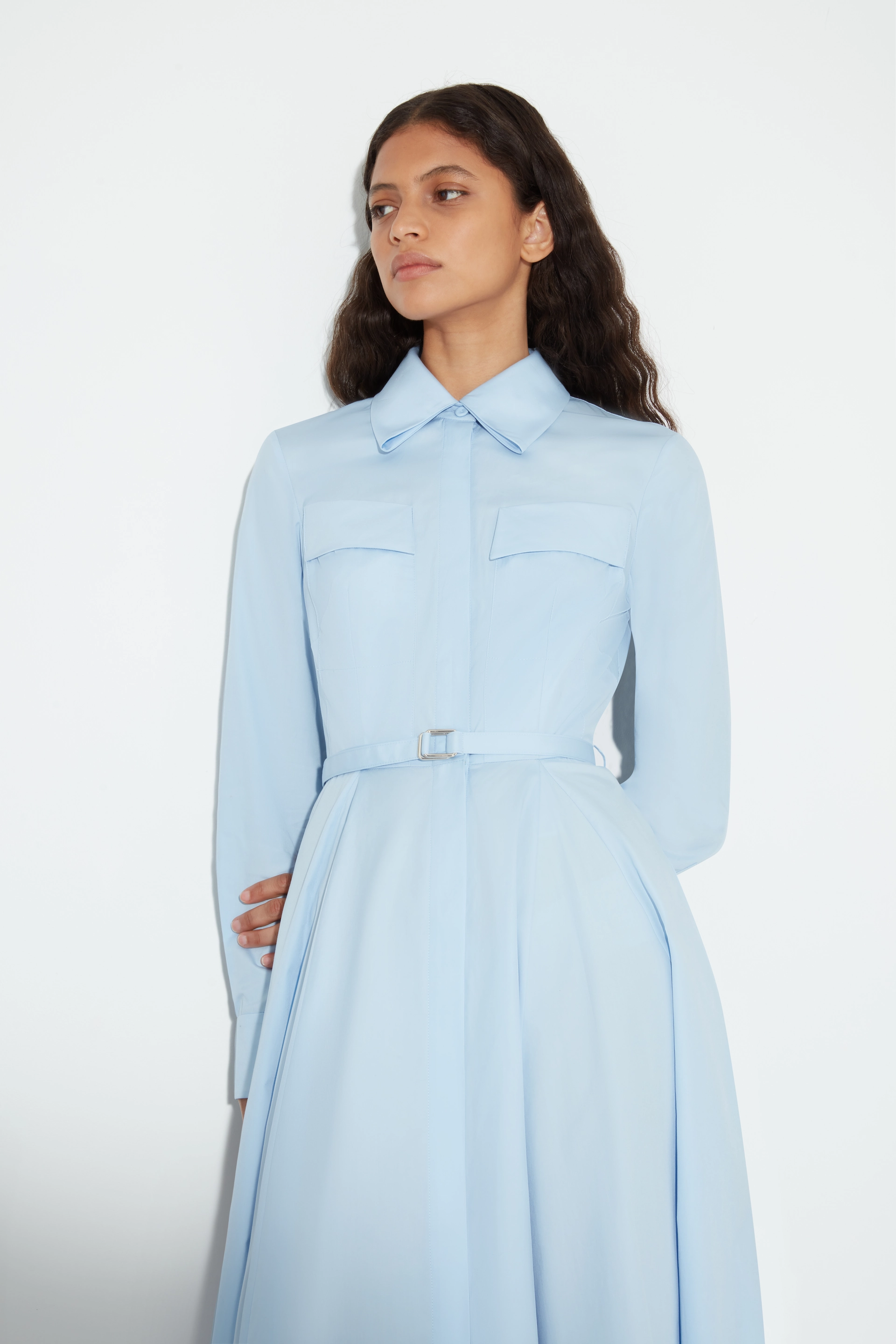 Woman with long wavy hair wearing a belted, light blue collared Emilia Wickstead dress with flap pockets.