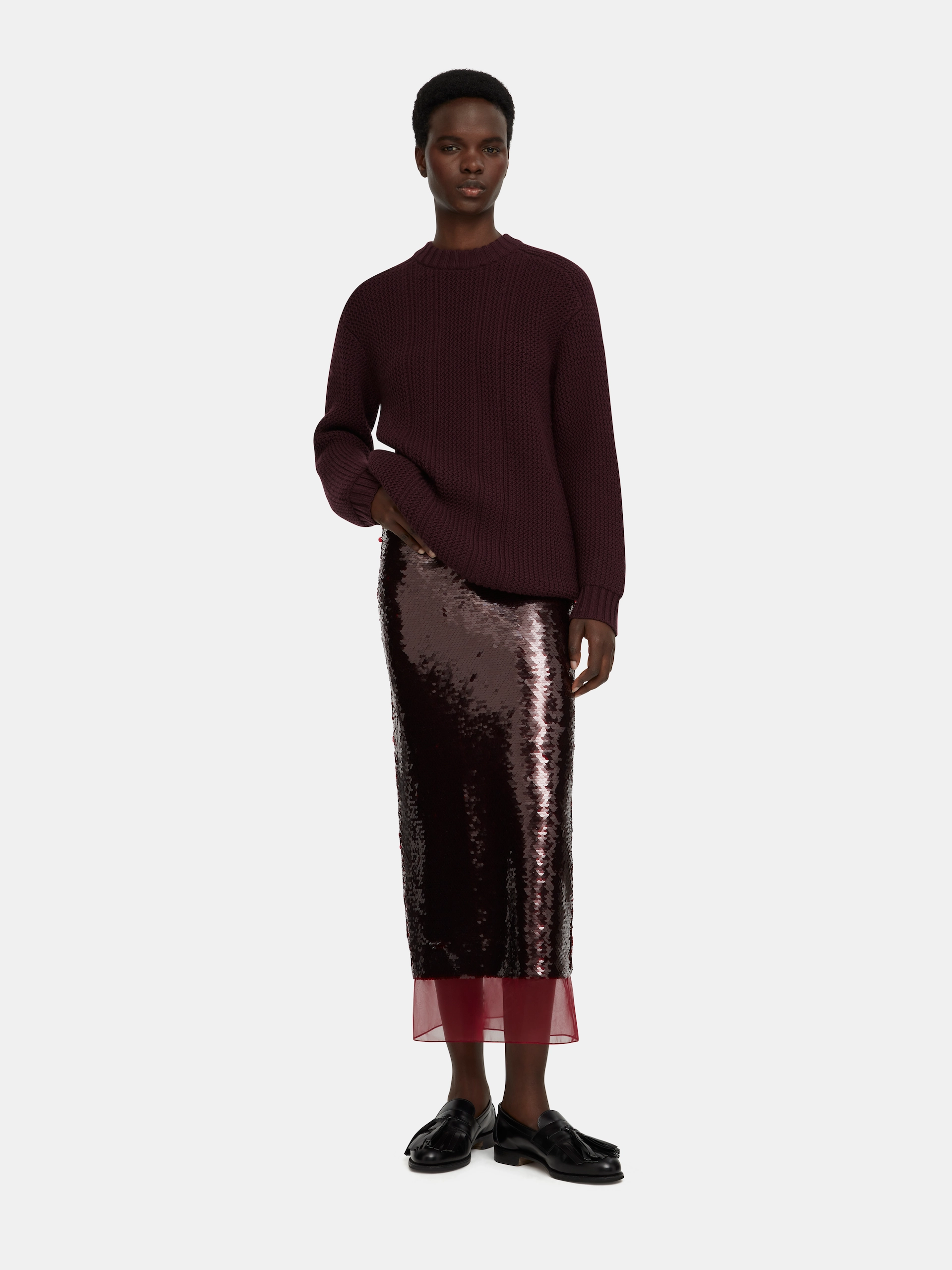 Model wearing a dark purple chunky knit Emilia Wickstead sweater and a sequined midi skirt with black loafers.