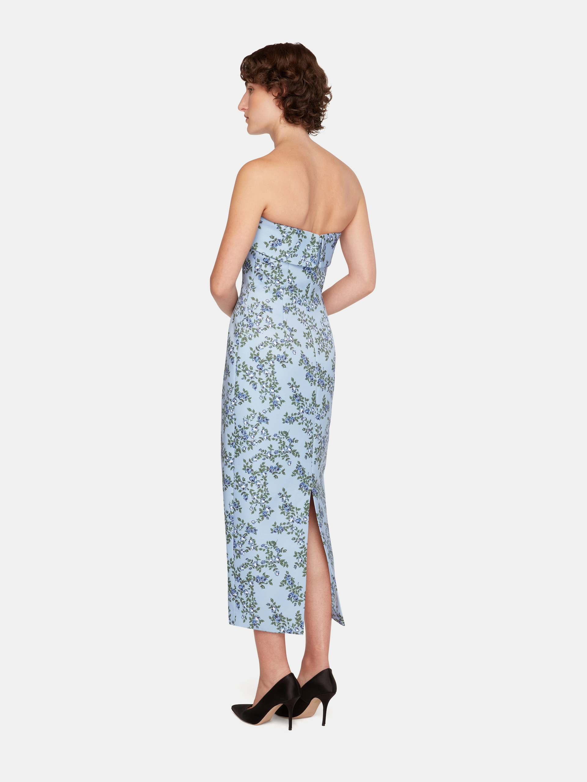 Woman in a strapless, light blue floral Emilia Wickstead dress with a back slit and black high heels, facing sideways.