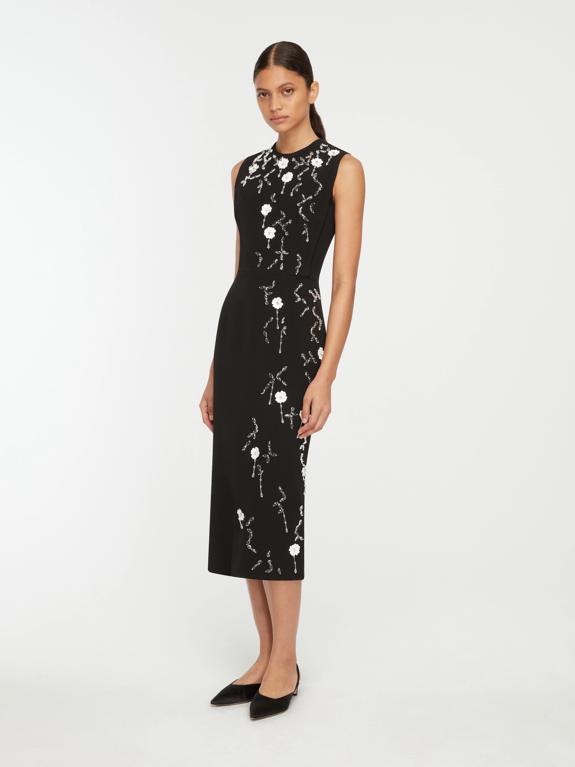 Model wearing a sleeveless black Emilia Wickstead midi dress with white floral embroidery and black pointed flats.