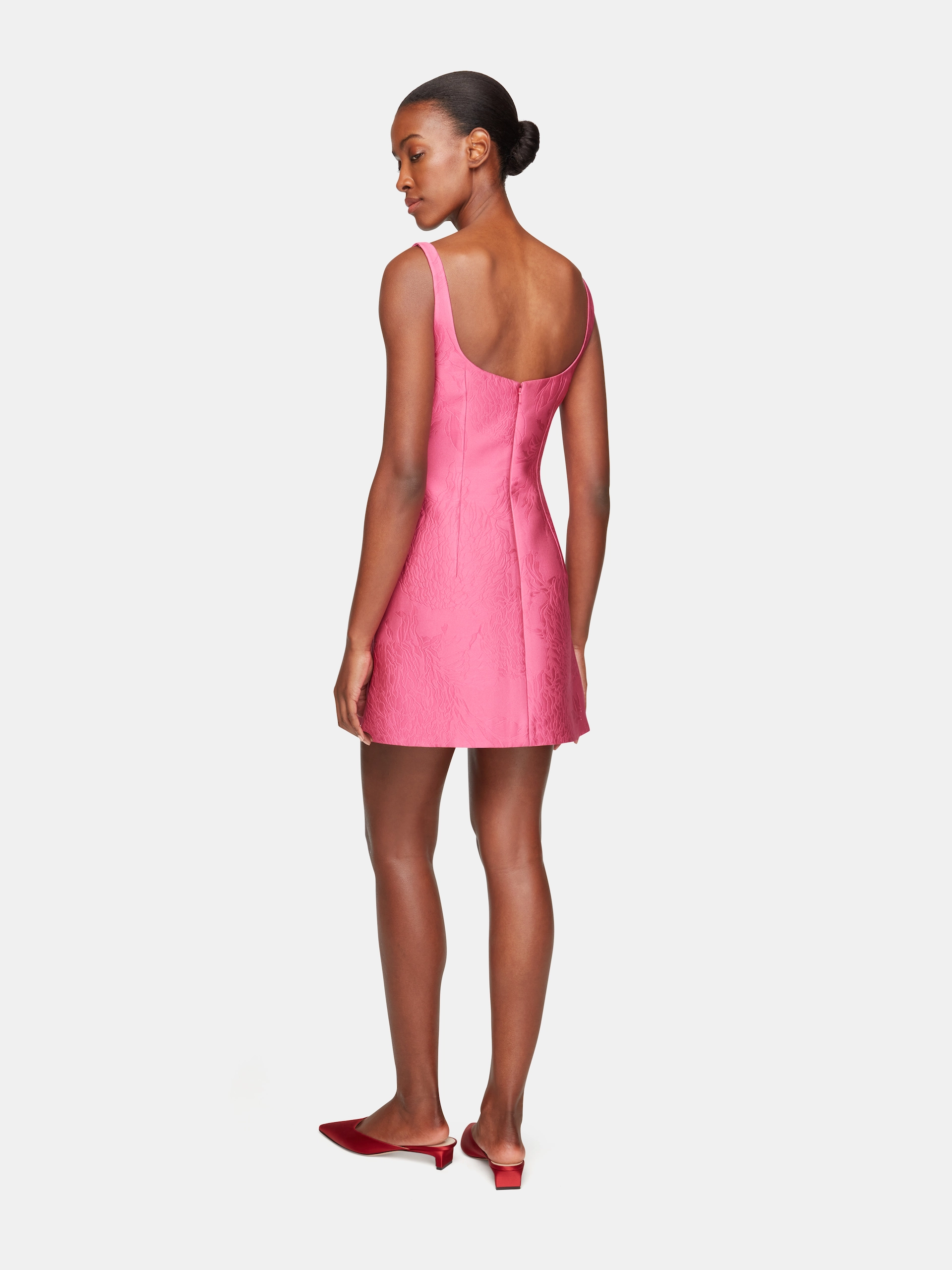 Woman wearing a short pink Emilia Wickstead sleeveless dress and red low-heeled mules, standing facing backward.