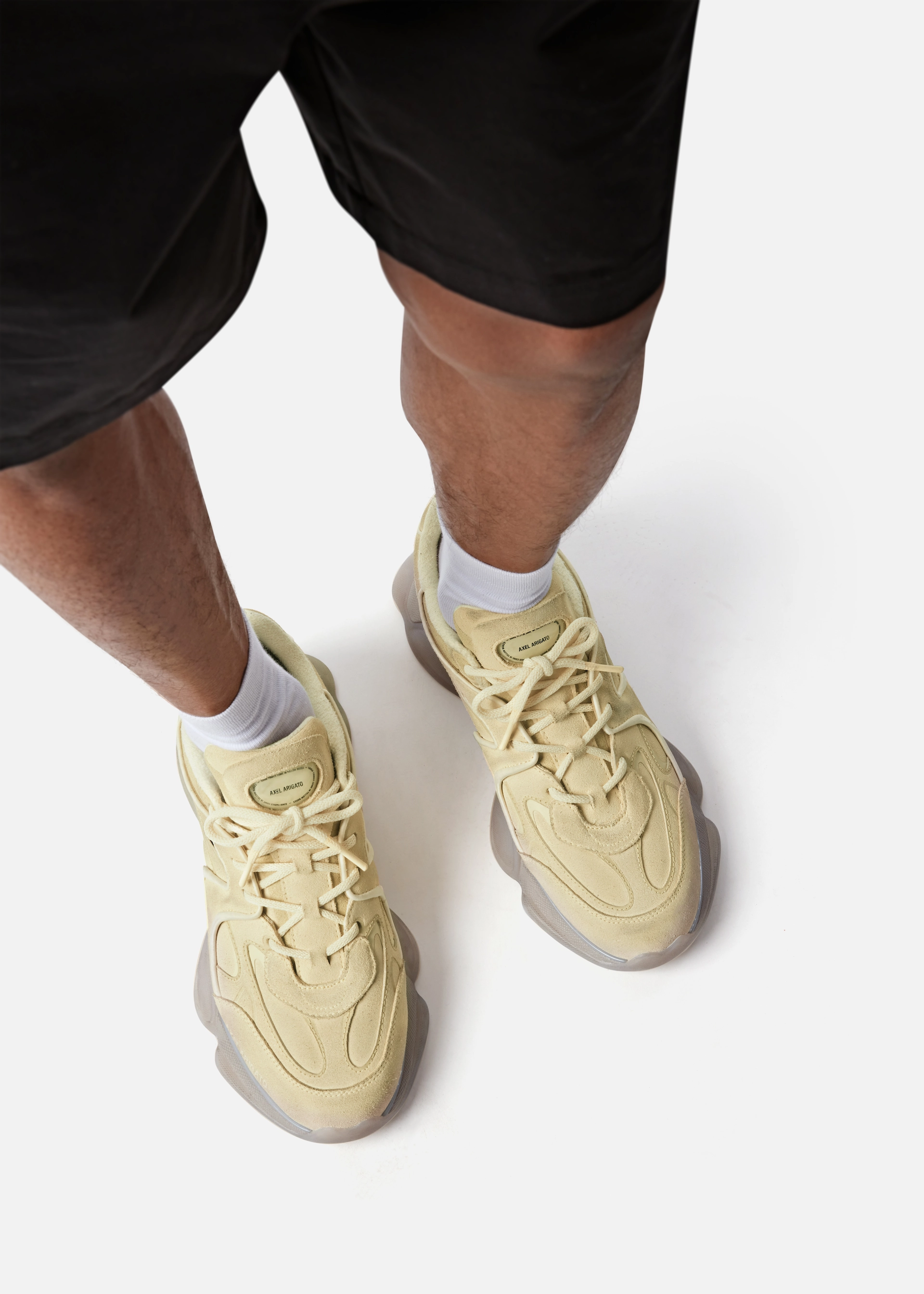 Person wearing cream Axel Arigato sneakers with white socks and black shorts on a white background.