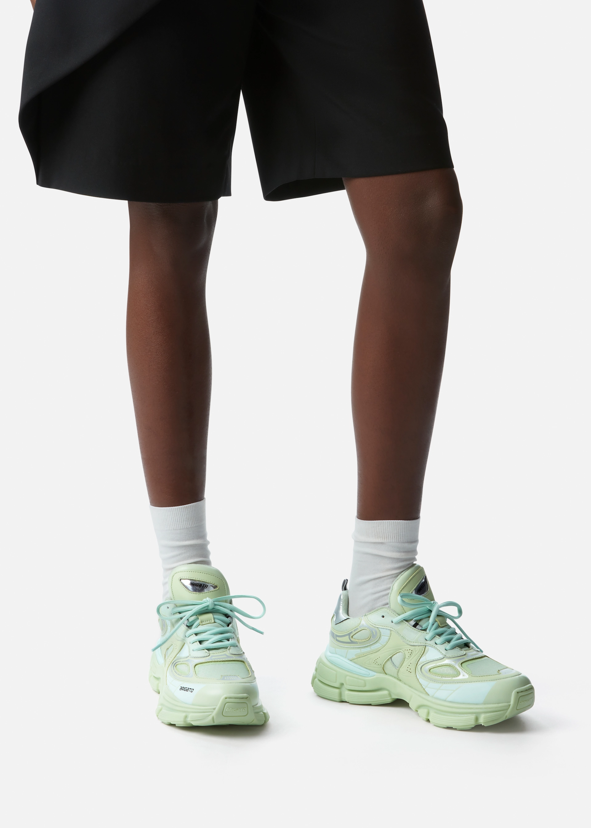 Person wearing light green chunky Axel Arigato sneakers with white socks and black shorts on a white background.