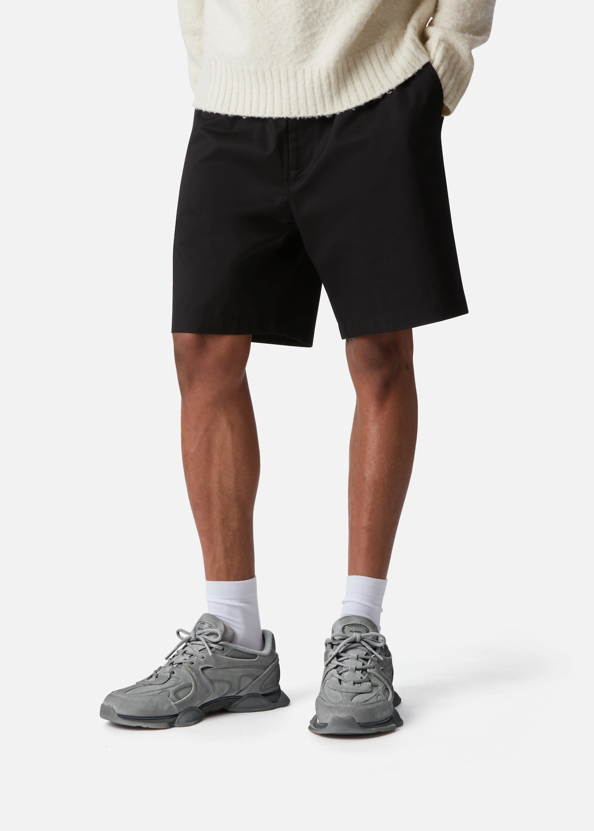 Person wearing black shorts, white socks, gray Axel Arigato sneakers, and a cream sweater with hands in pockets.