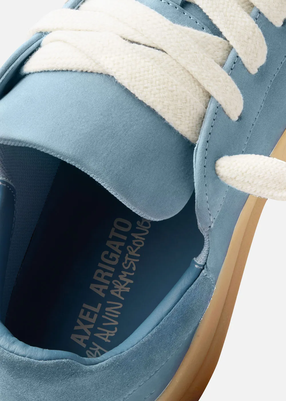 Close-up of a blue suede Axel Arigato sneaker with cream laces and a gum sole, showing the brand name inside.