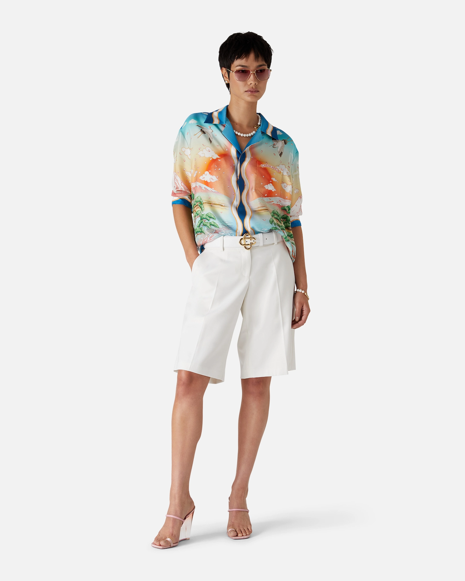 Model in colorful Casablanca scenic shirt, white belted shorts, pearl jewelry, pink heeled sandals, and sunglasses.
