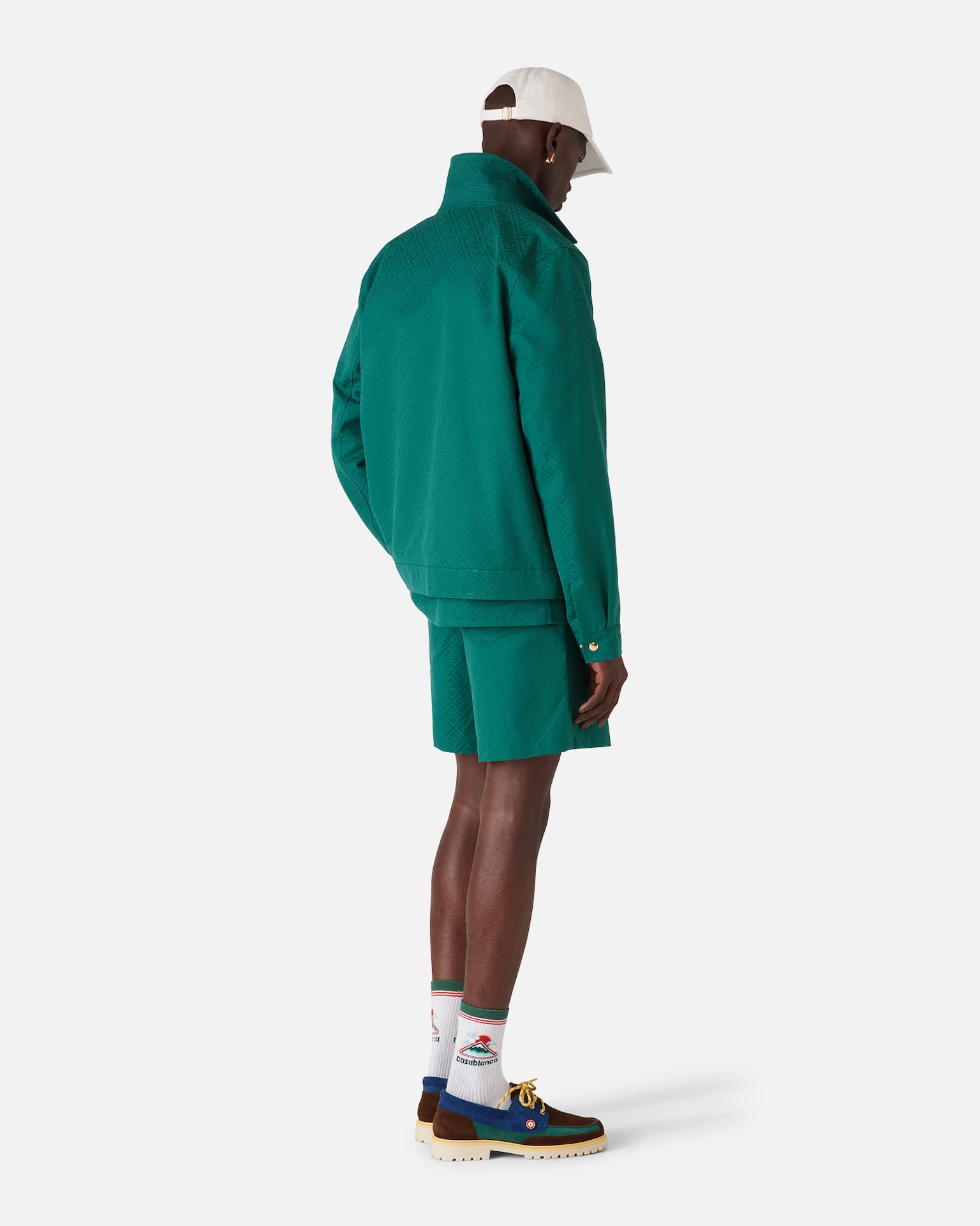 Man wearing a teal patterned Casablanca jacket and shorts, white cap, white Casablanca logo socks, and color-block shoes.