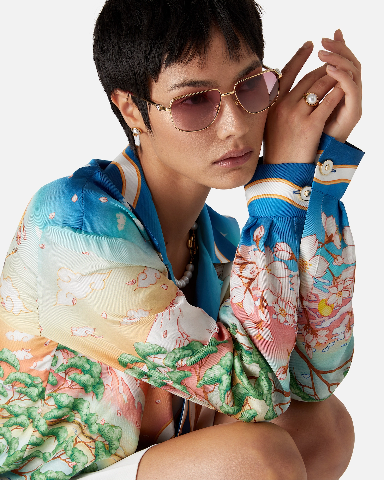 Person wearing a colorful Casablanca floral-patterned shirt, pink-tinted sunglasses, pearl earrings, and ring.