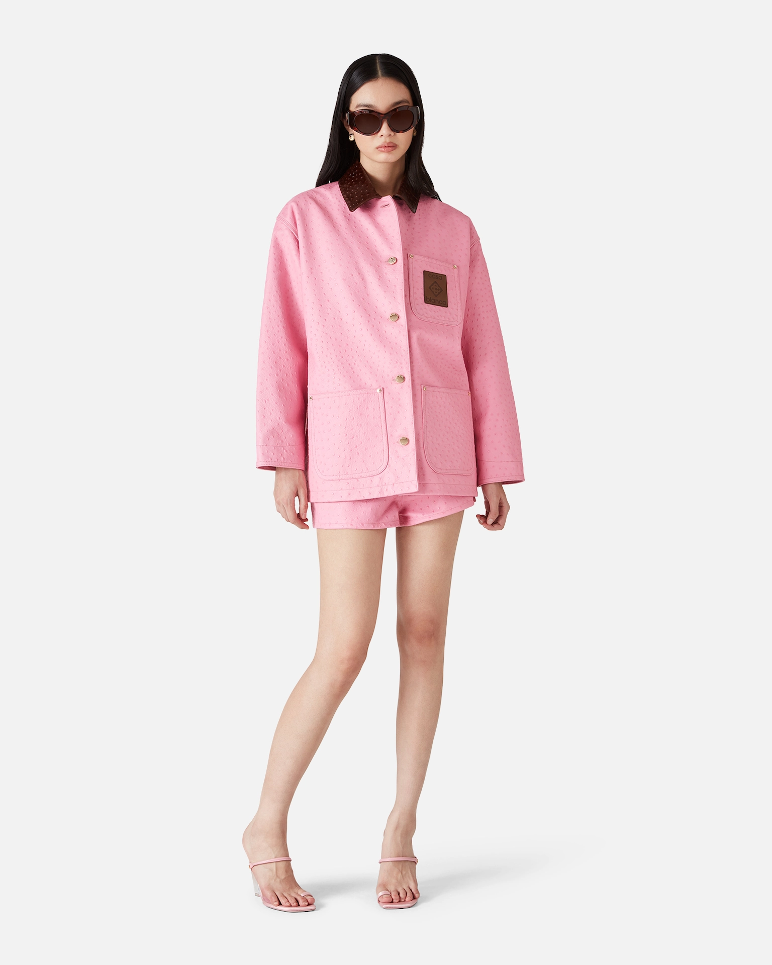 Woman wearing pink textured Casablanca jacket with brown collar, matching shorts, sunglasses, and pink heeled sandals.