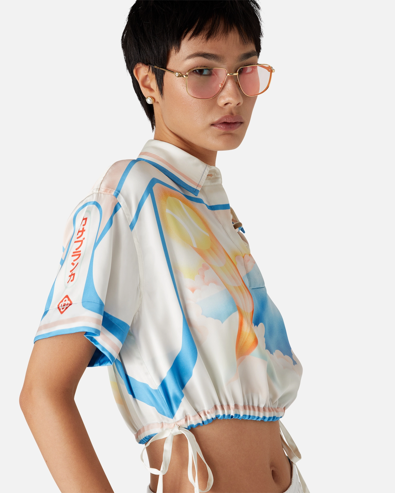 Woman with short black hair wearing pink-tinted Casablanca glasses and a colorful cropped shirt with blue and orange patterns.