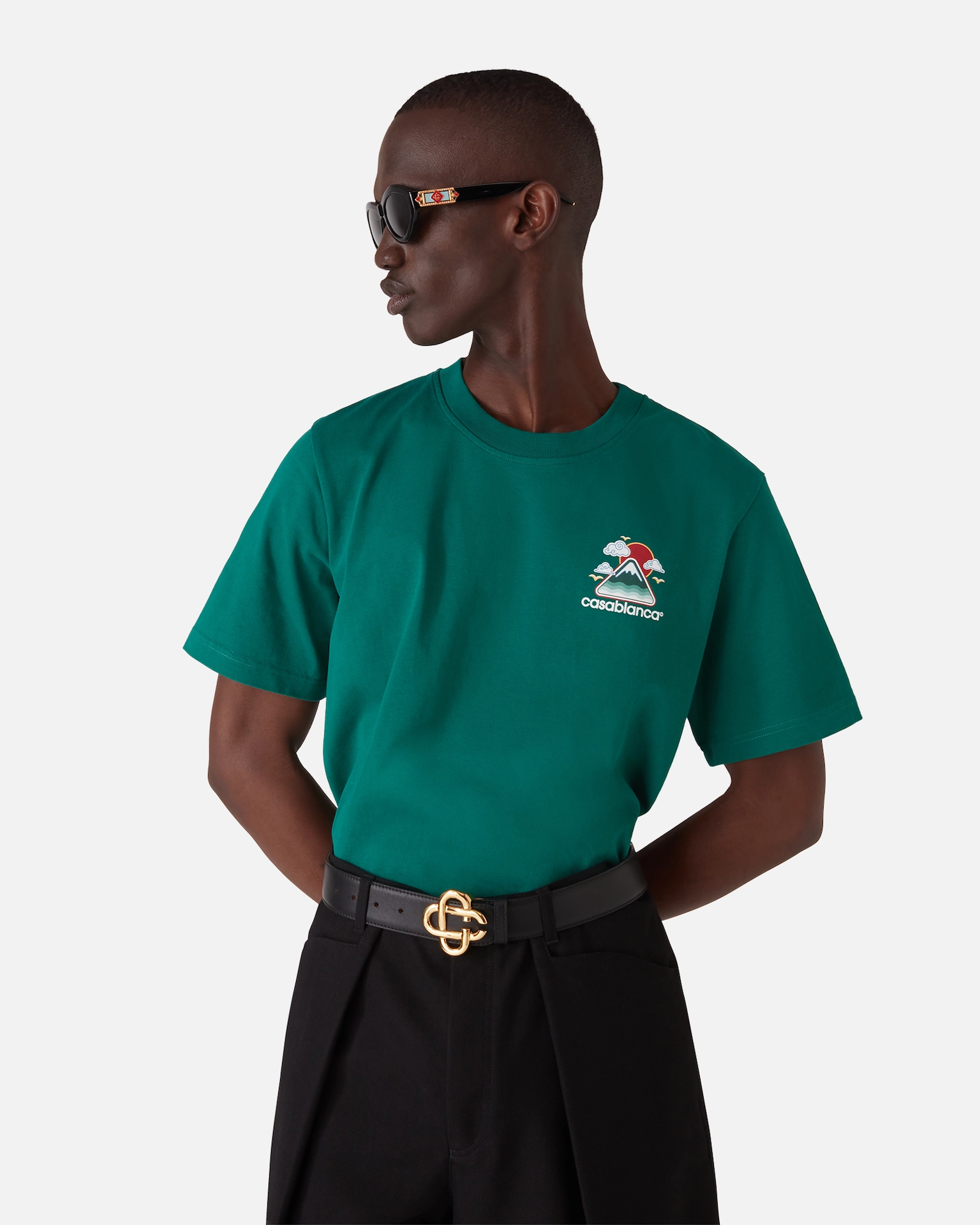 Man wearing green Casablanca t-shirt with mountain logo and black pants with gold-buckled belt.