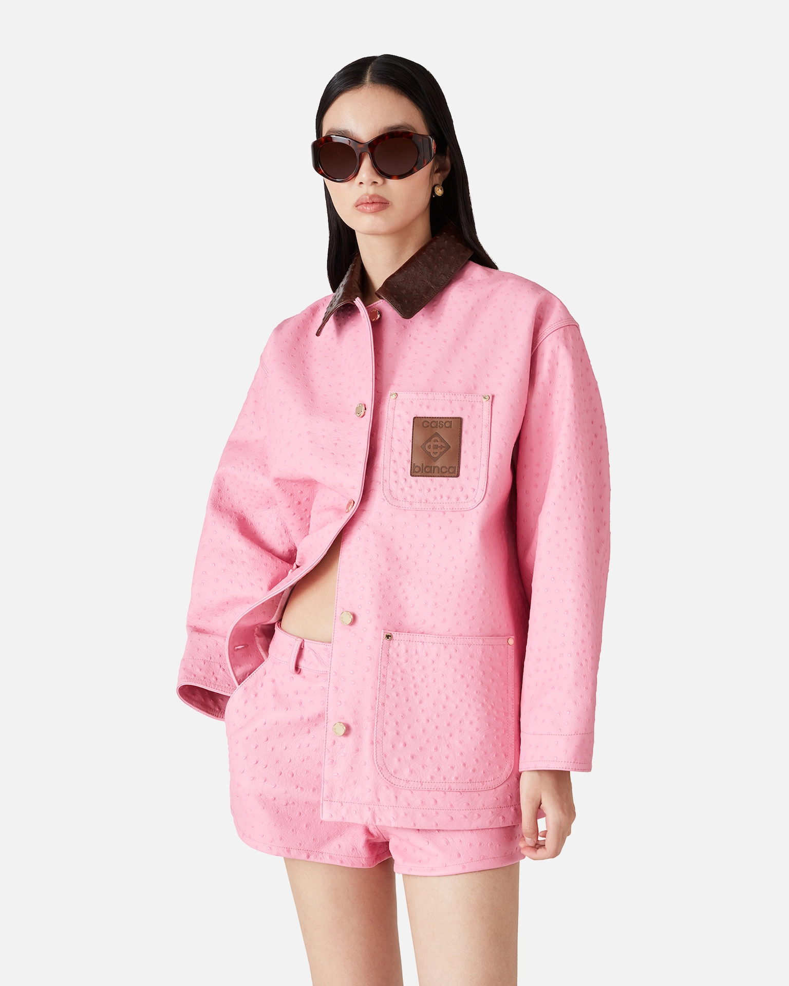 Woman wearing textured pink Casablanca jacket and matching shorts with brown collar and patch, posing with sunglasses.