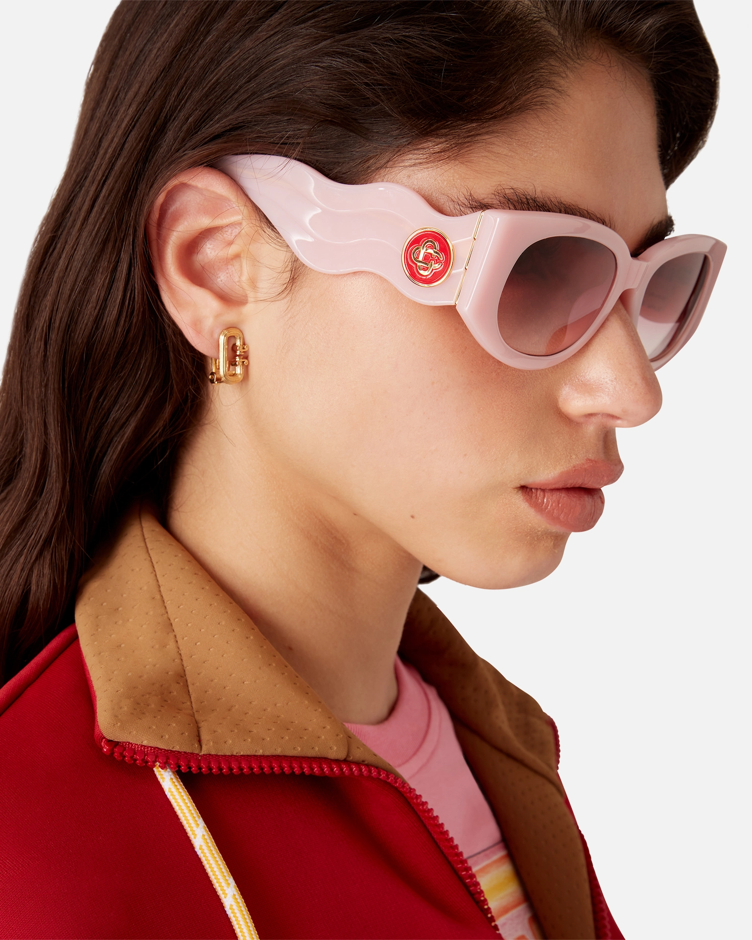 Side view of woman wearing pink Casablanca winged sunglasses and gold earrings in red jacket with brown collar.