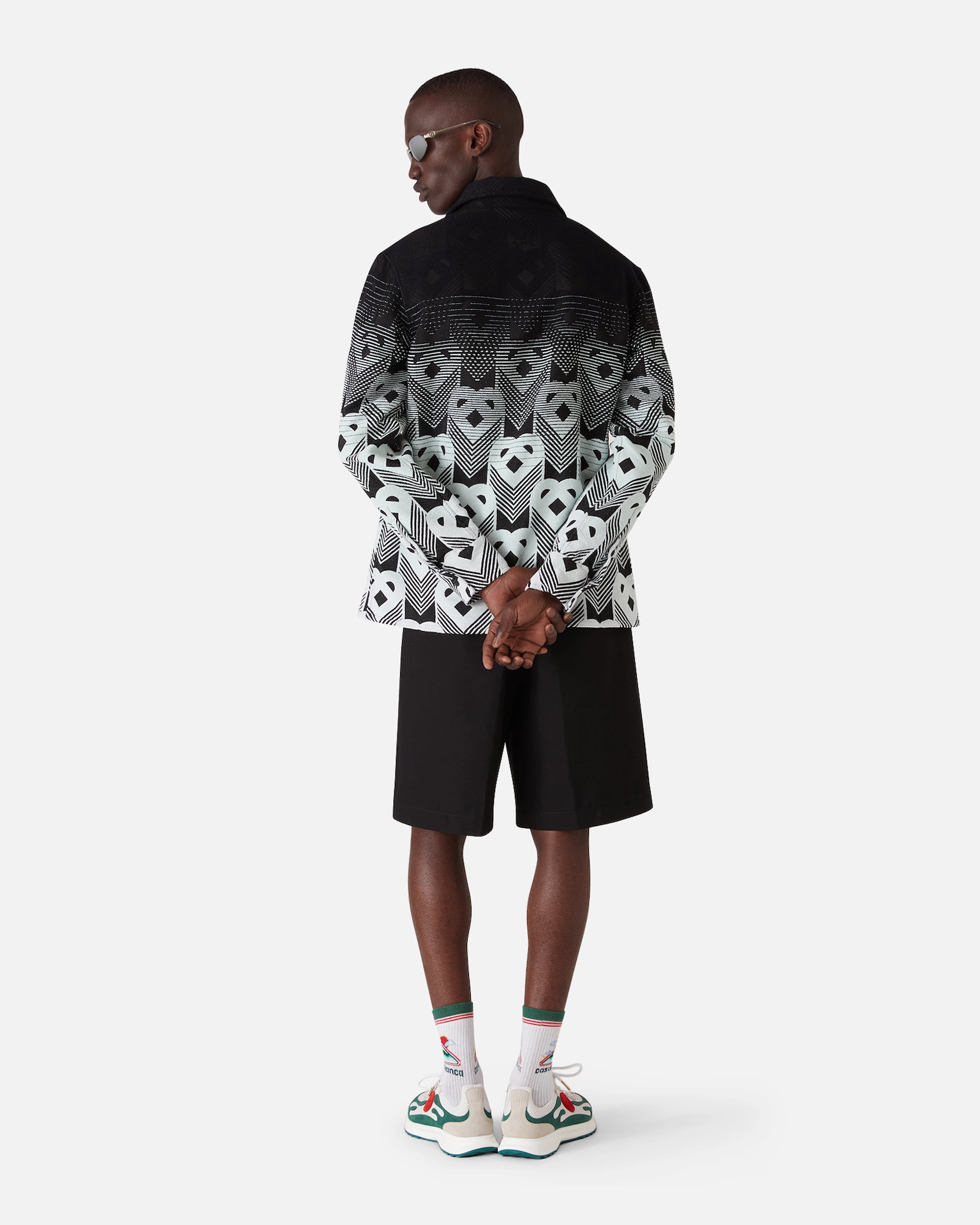 Man wearing a patterned black Casablanca and white jacket, black shorts, white socks, and multicolor sneakers, facing away.