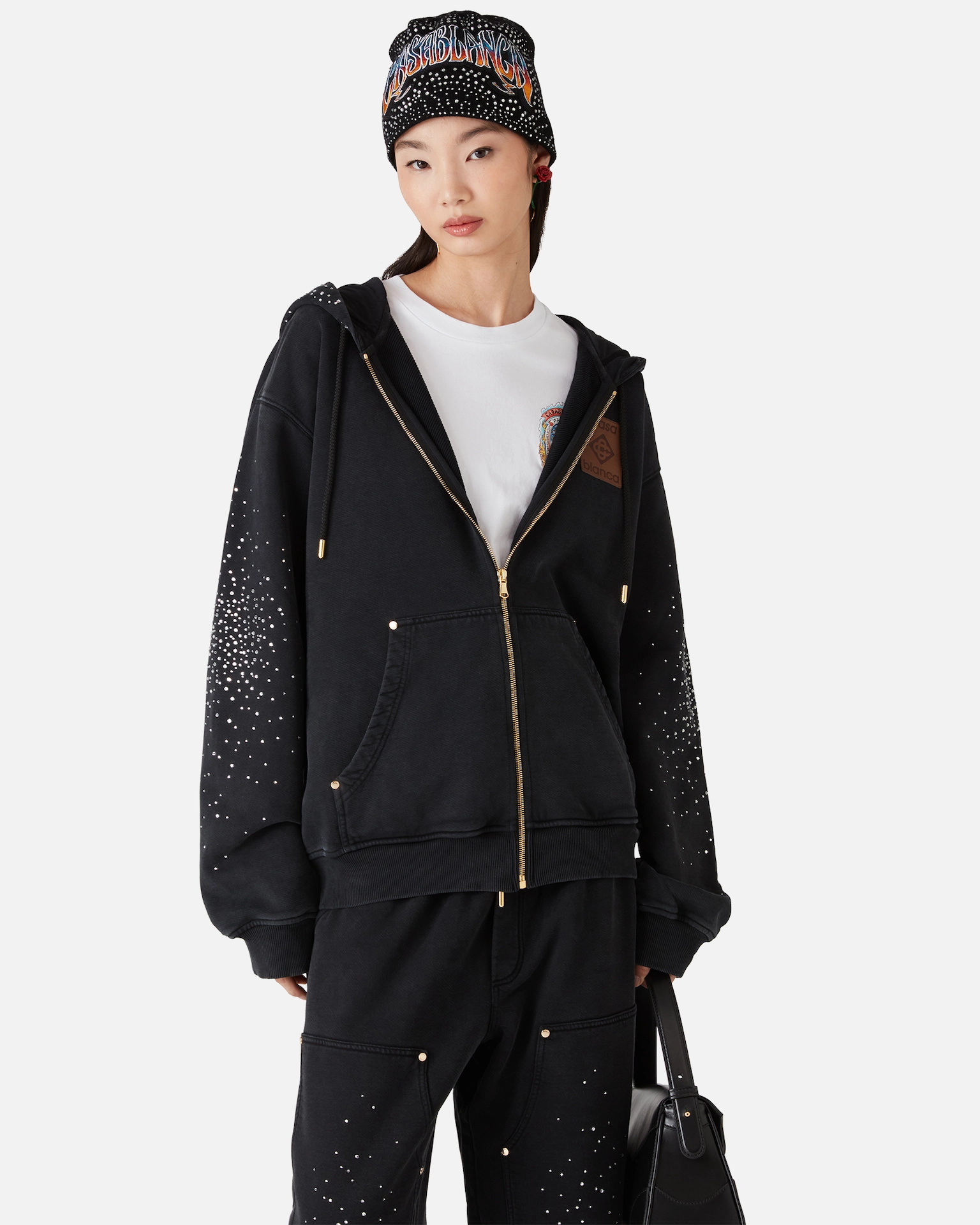 Woman wearing black Casablanca zip-up hoodie and pants with white speckles, a printed beanie, and holding a black bag.