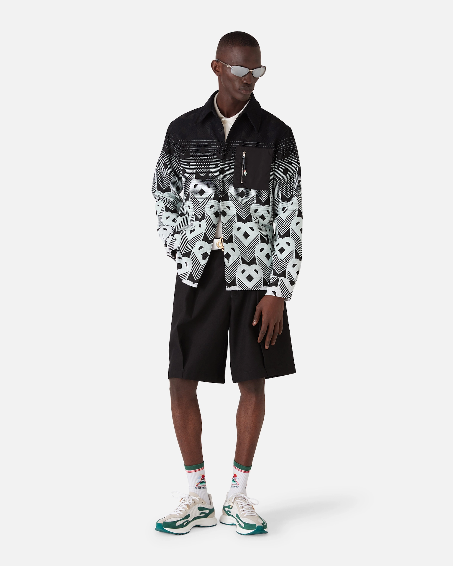 Man wearing patterned Casablanca jacket, black shorts, white socks with logo, and green-white sneakers with sunglasses.