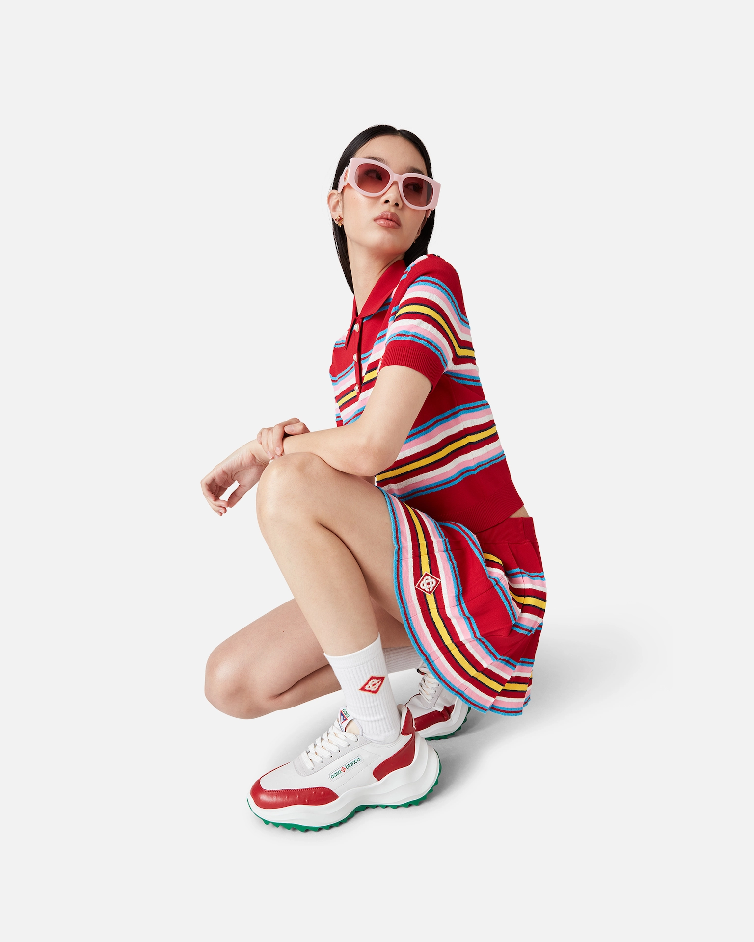 Woman crouching in red striped Casablanca dress, white socks, white sneakers with red accents, and pink sunglasses.