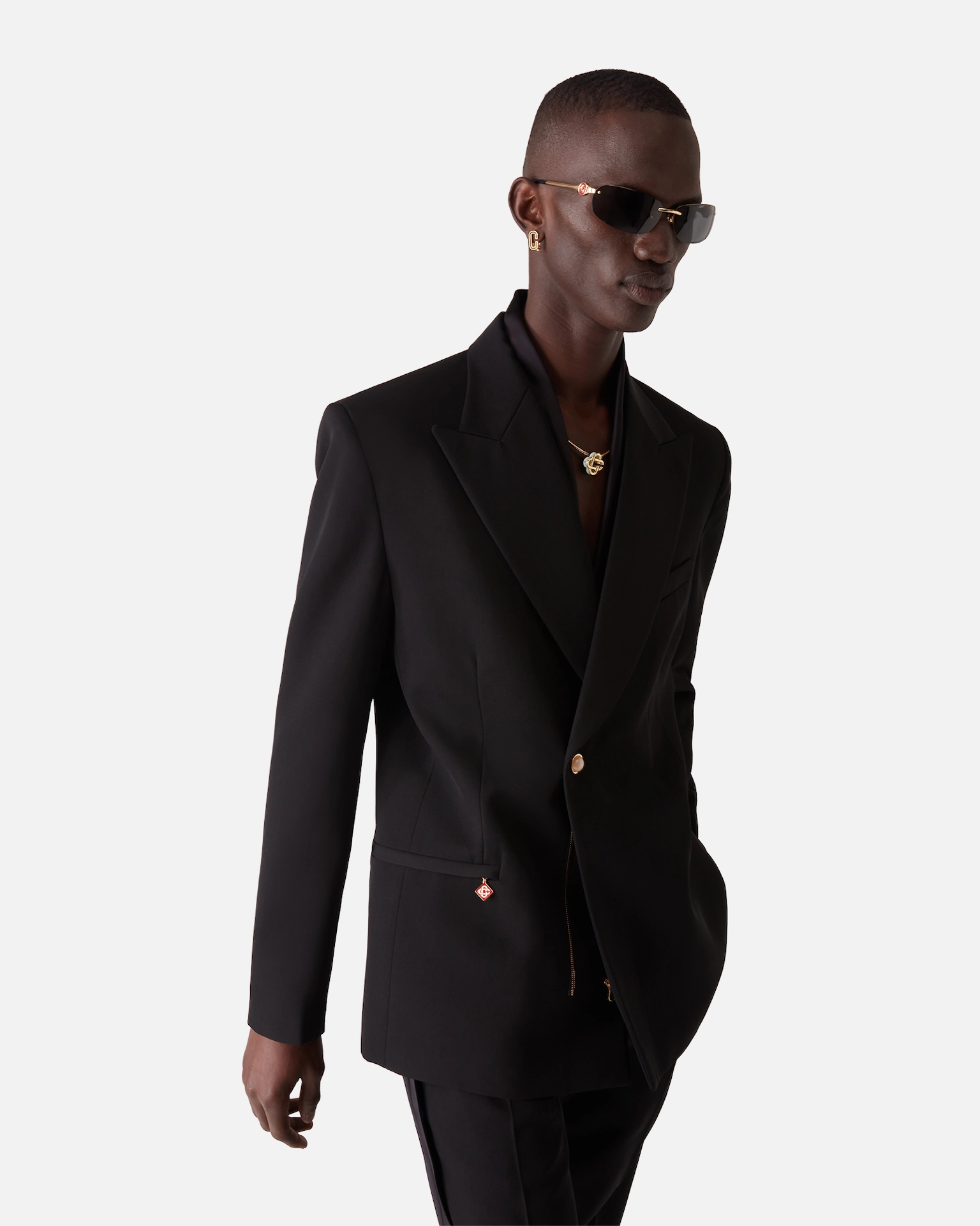 Man wearing black Casablanca suit, gold necklace, gold earrings, and dark sunglasses on white background.