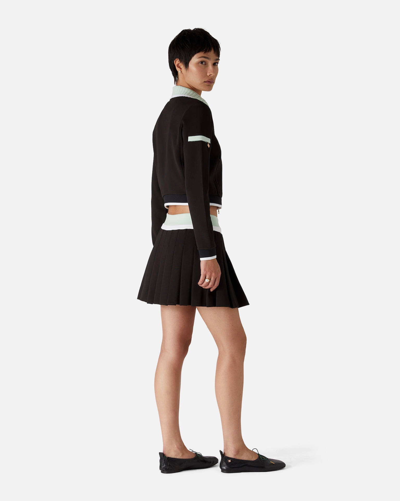 Woman in black Casablanca pleated skirt and cropped jacket with light blue trim, standing sideways on white background.