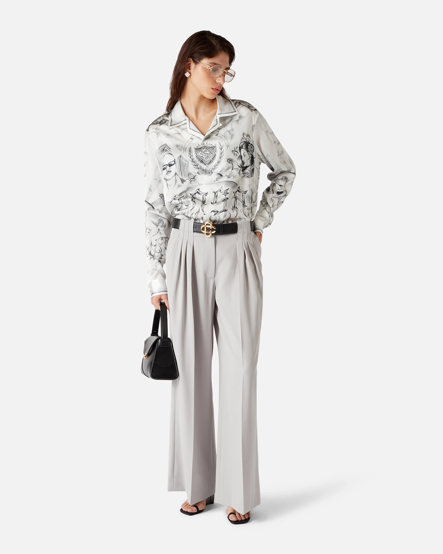 Woman wearing a Casablanca patterned long-sleeve shirt, light gray wide-leg pants, black belt, sandals, and holding a black handbag.