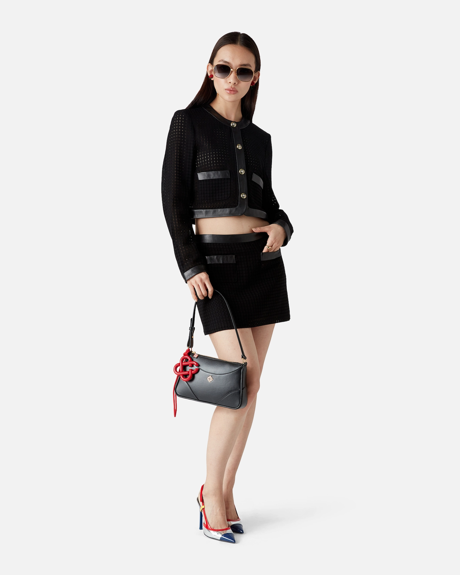 Woman wearing black Casablanca cropped jacket and skirt, holding black handbag with red charm, in sunglasses.