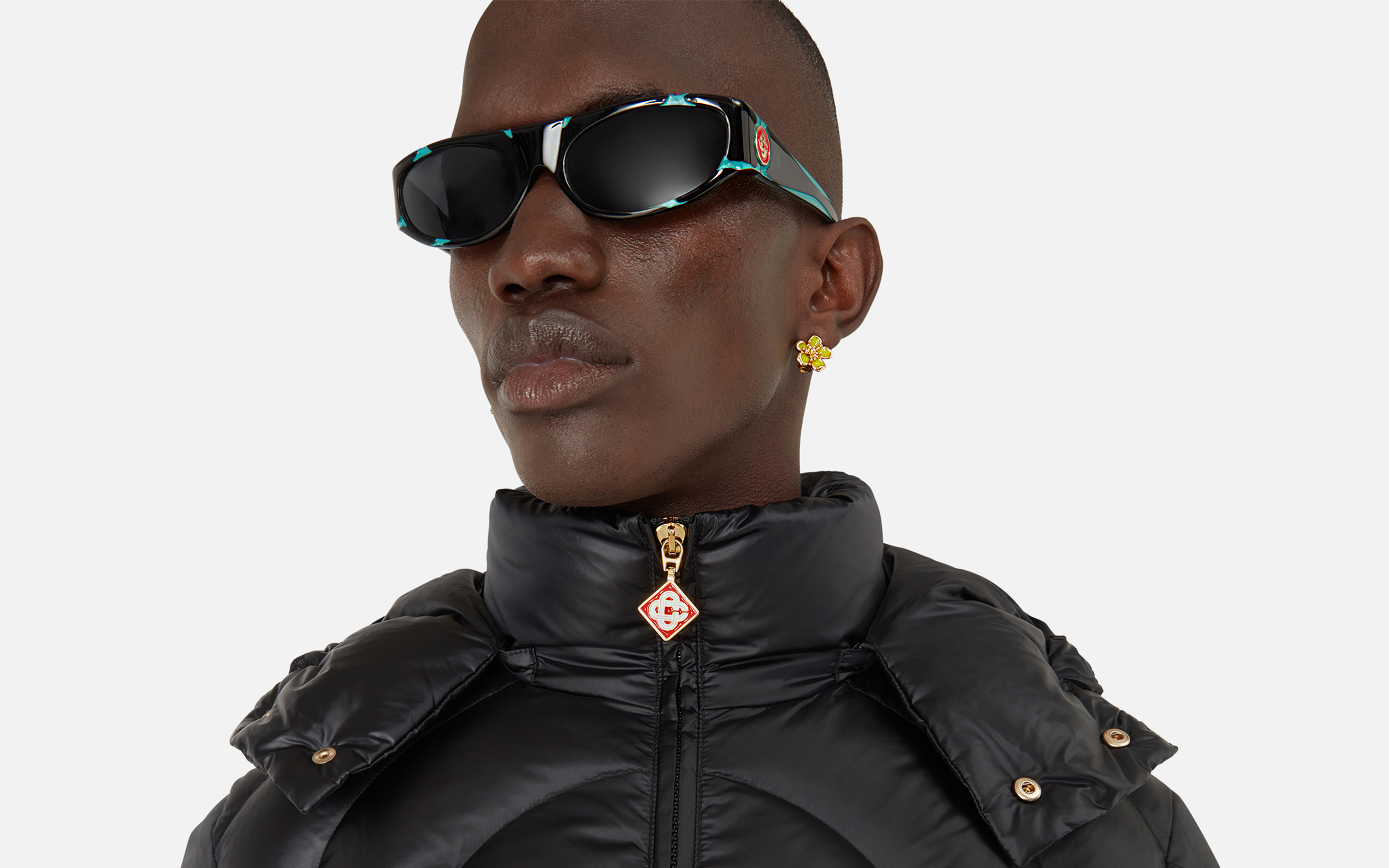 Close-up of a person wearing black Casablanca sunglasses, a black puffer jacket, and a yellow flower-shaped earring.