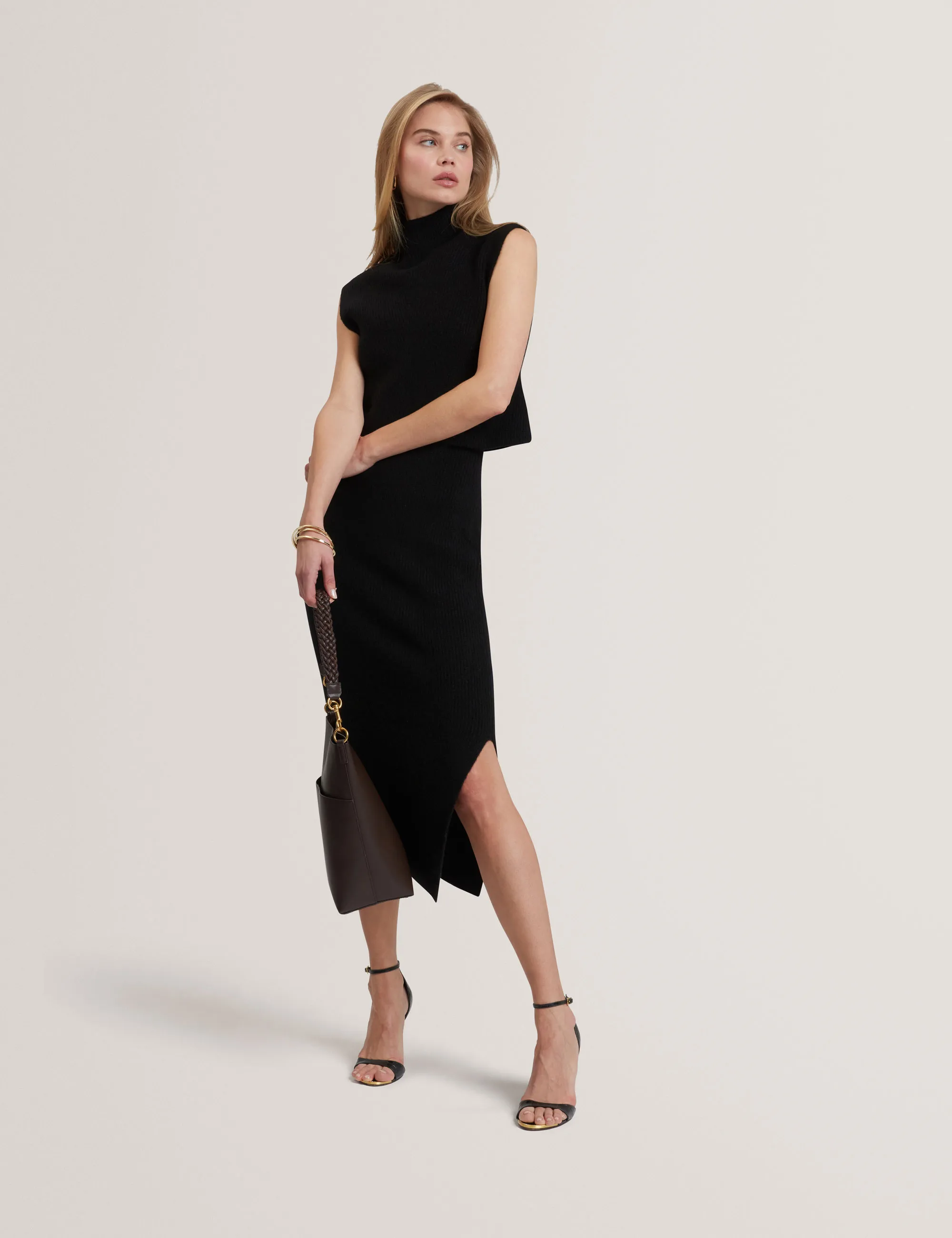 Woman wearing a black sleeveless Ted Baker turtleneck top and matching asymmetric midi skirt holding a brown purse.
