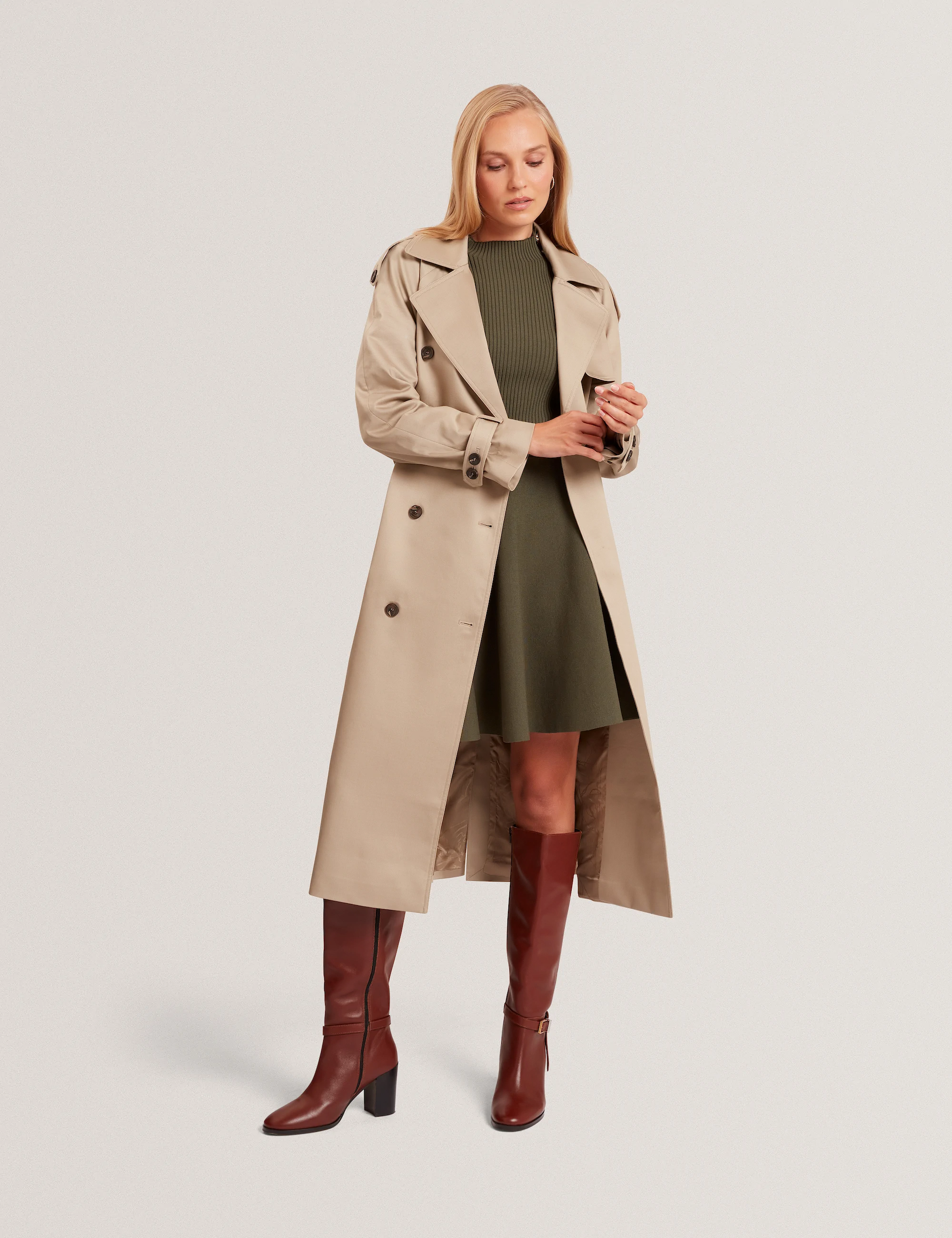 Woman wearing a beige Ted Baker  trench coat over an olive dress and knee-high brown leather boots.