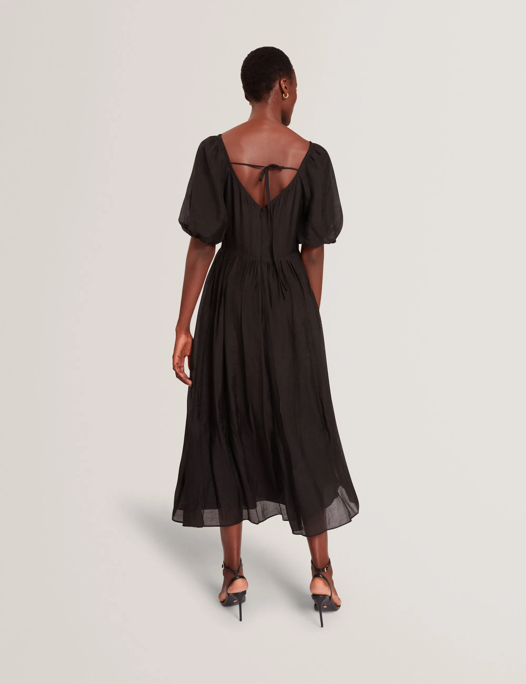 Woman wearing a black Ted Baker  midi dress with puff sleeves and a deep back tied with a thin strap.
