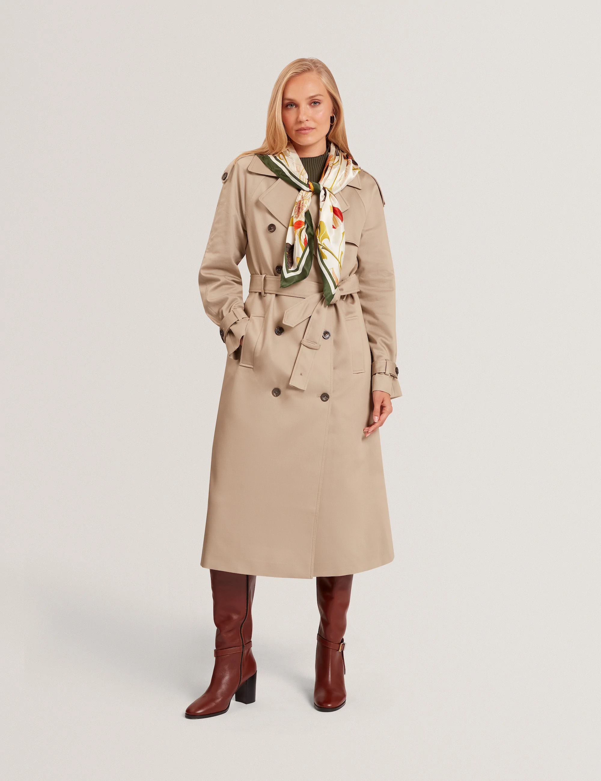 Woman wearing a beige Ted Baker  trench coat, patterned scarf, and brown knee-high boots standing against a plain background.