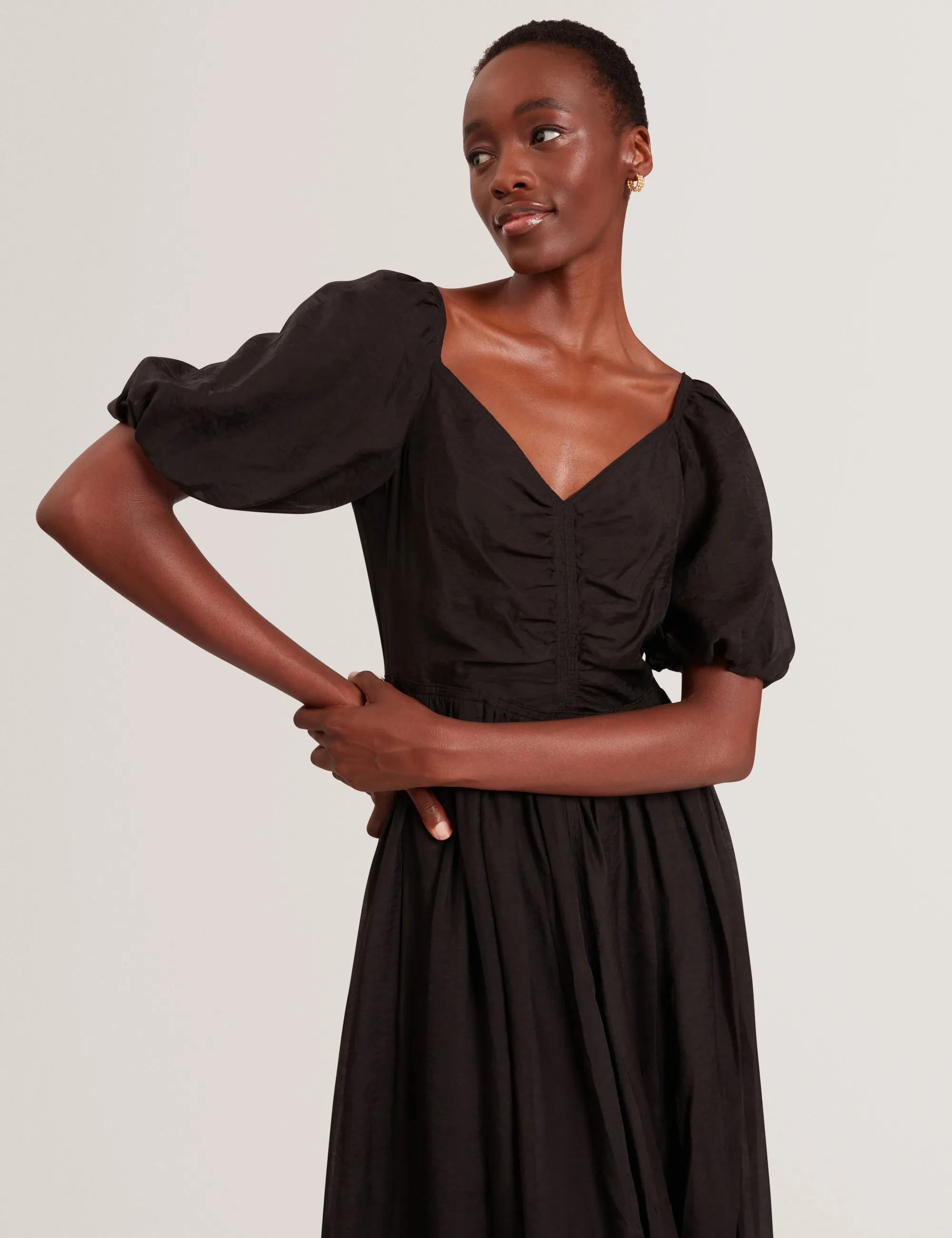 Woman wearing a black Ted Baker  dress with puff sleeves and ruched front posing against a light background.