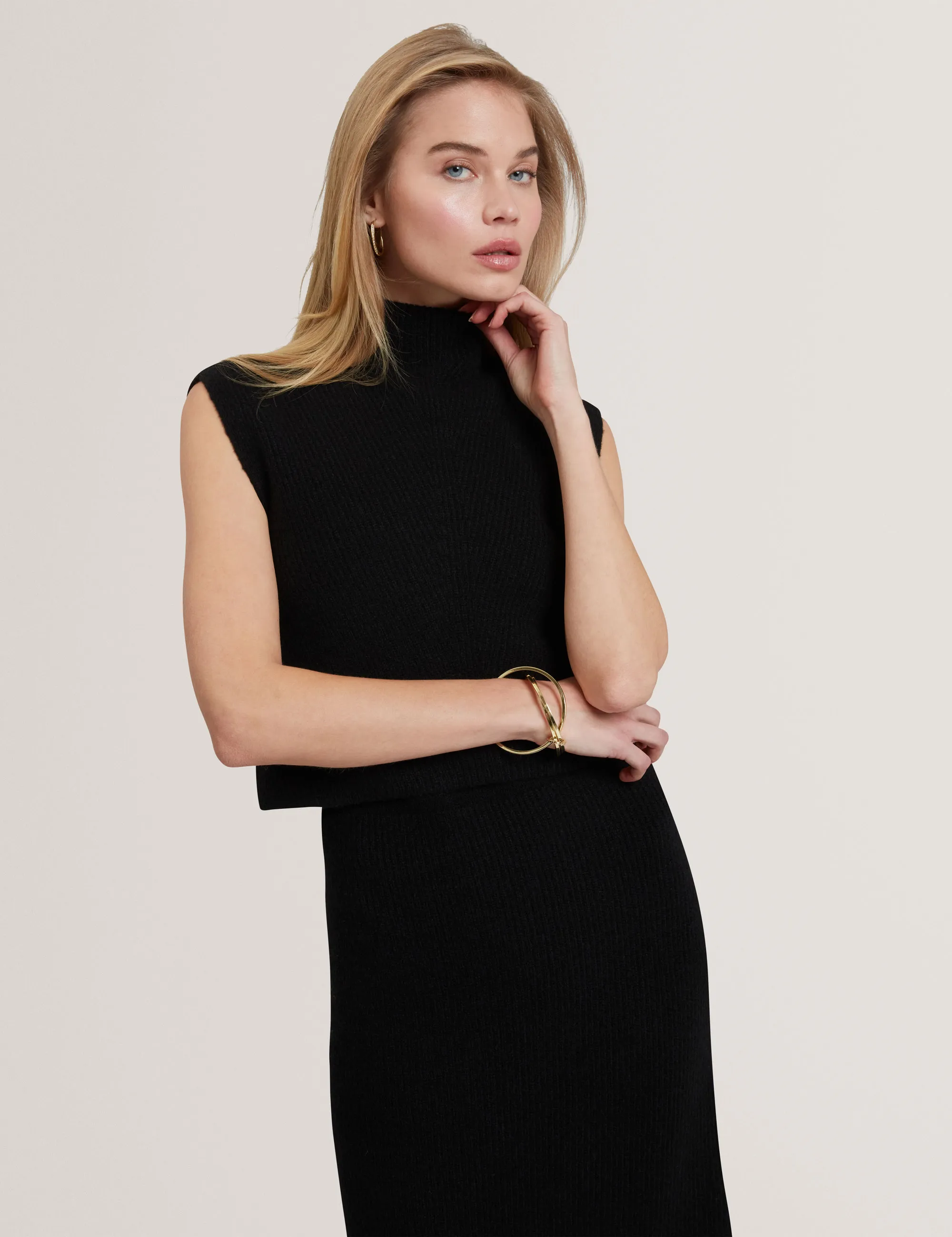 Blonde woman wearing sleeveless black Ted Baker  turtleneck and skirt, posing with one hand near face and gold bracelet.