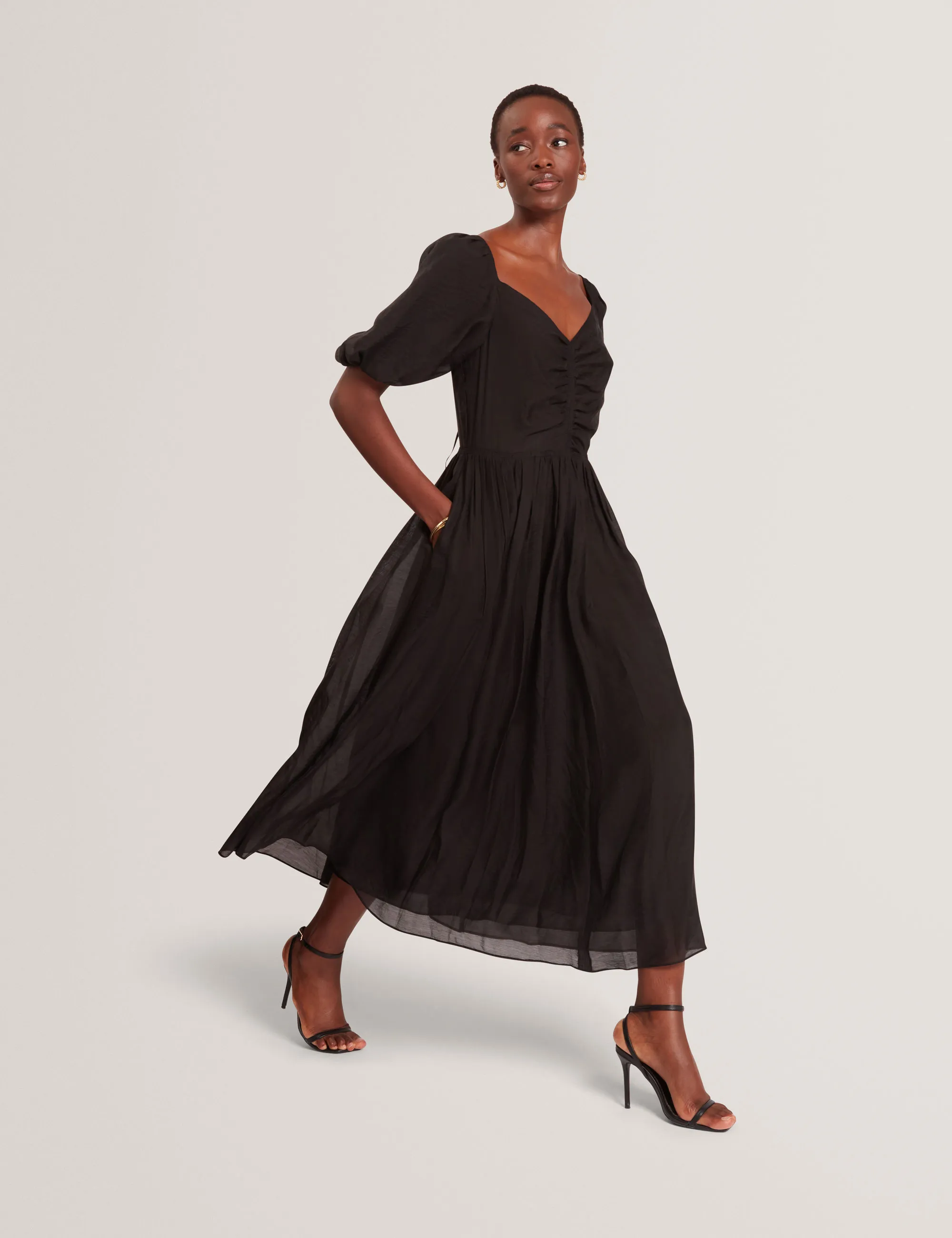Woman walking in a flowing black Ted Baker midi dress with puffed sleeves and black high heels.