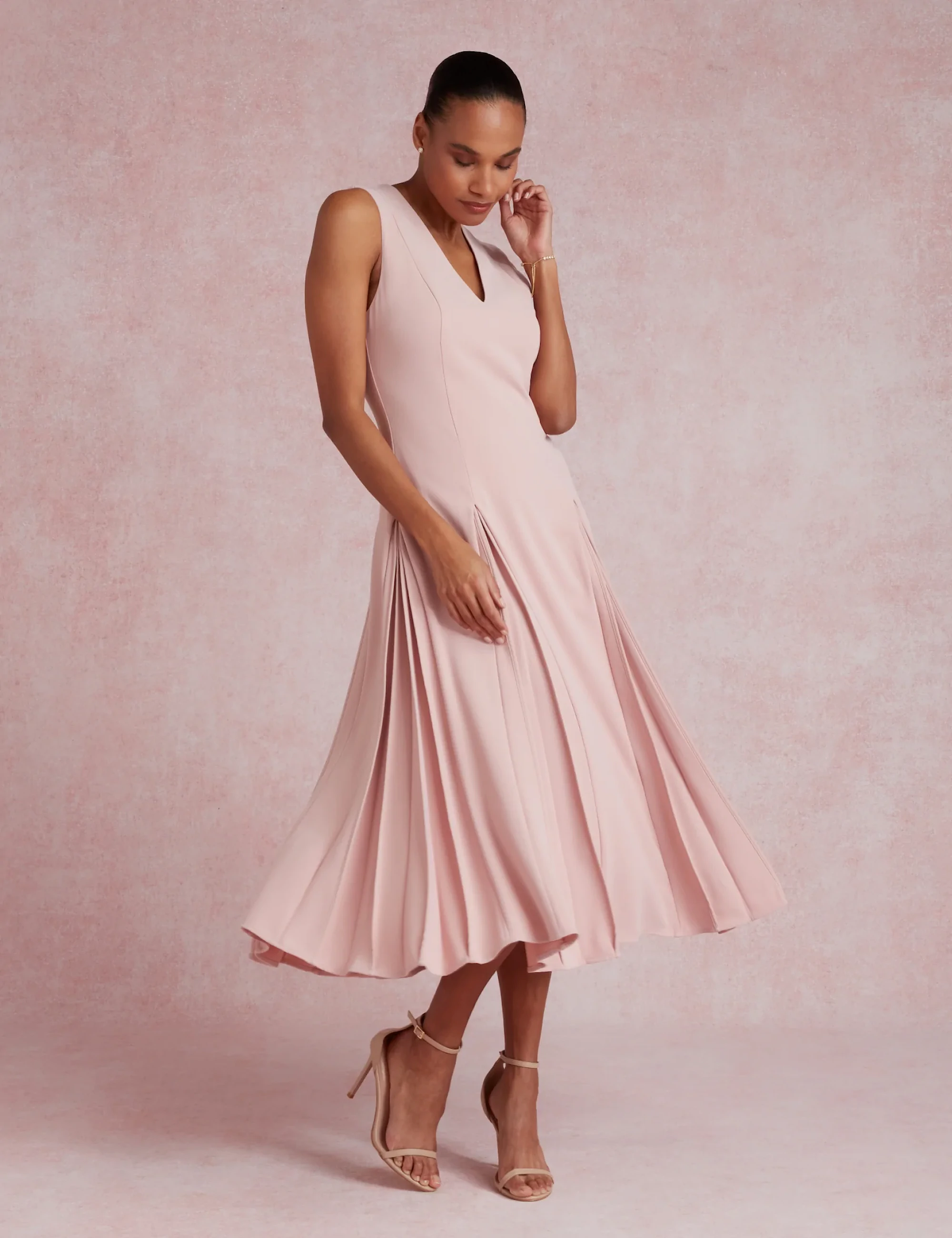 Woman wearing a sleeveless Ted Baker pale pink pleated midi dress with nude high heel sandals.