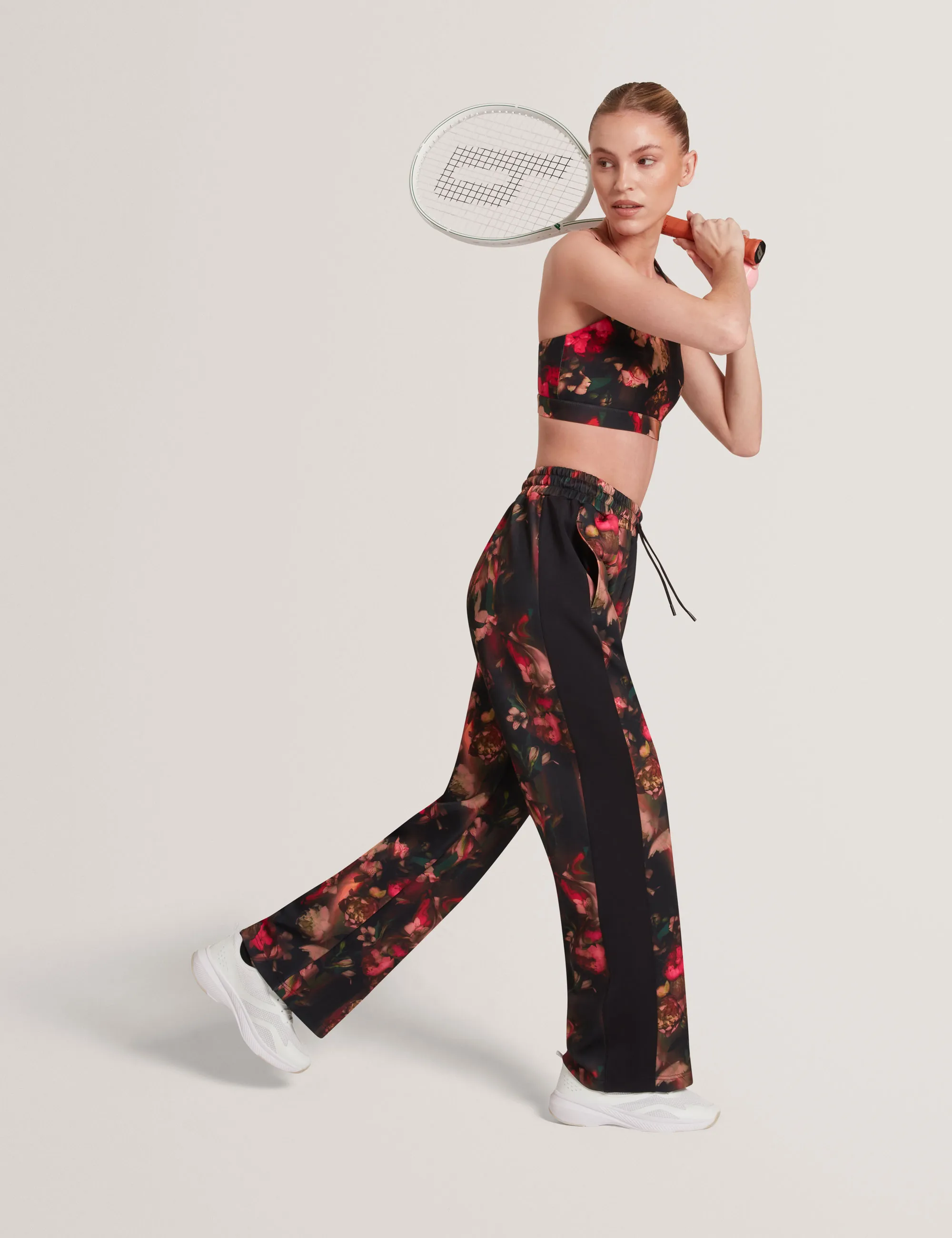 Woman in floral Ted Baker sports bra and pants holding a tennis racket in a ready-to-swing pose.
